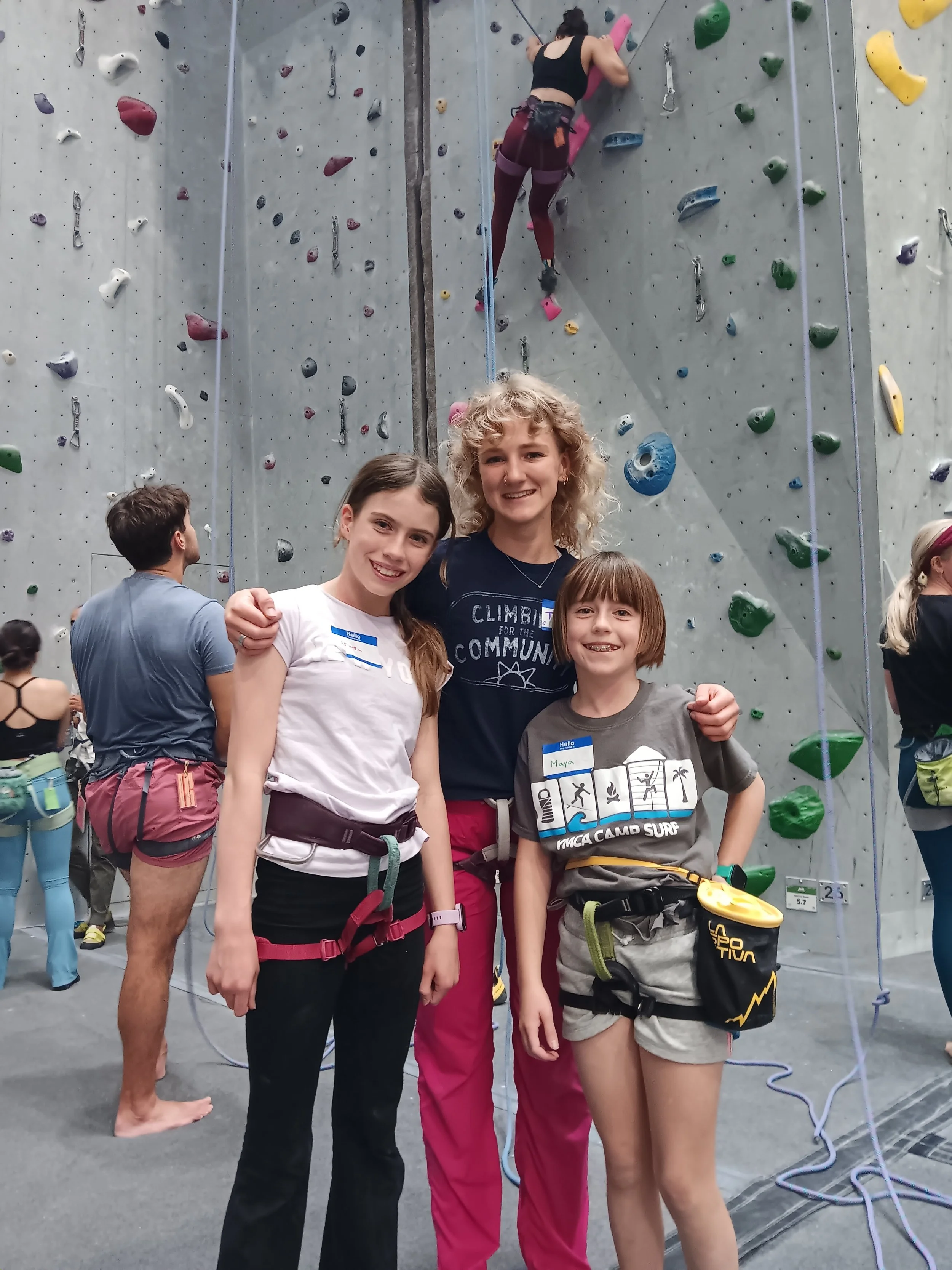 Three young girls with climbing gear smiling in front of an indoor climbing wall, with a woman climbing and other climbers in the background.