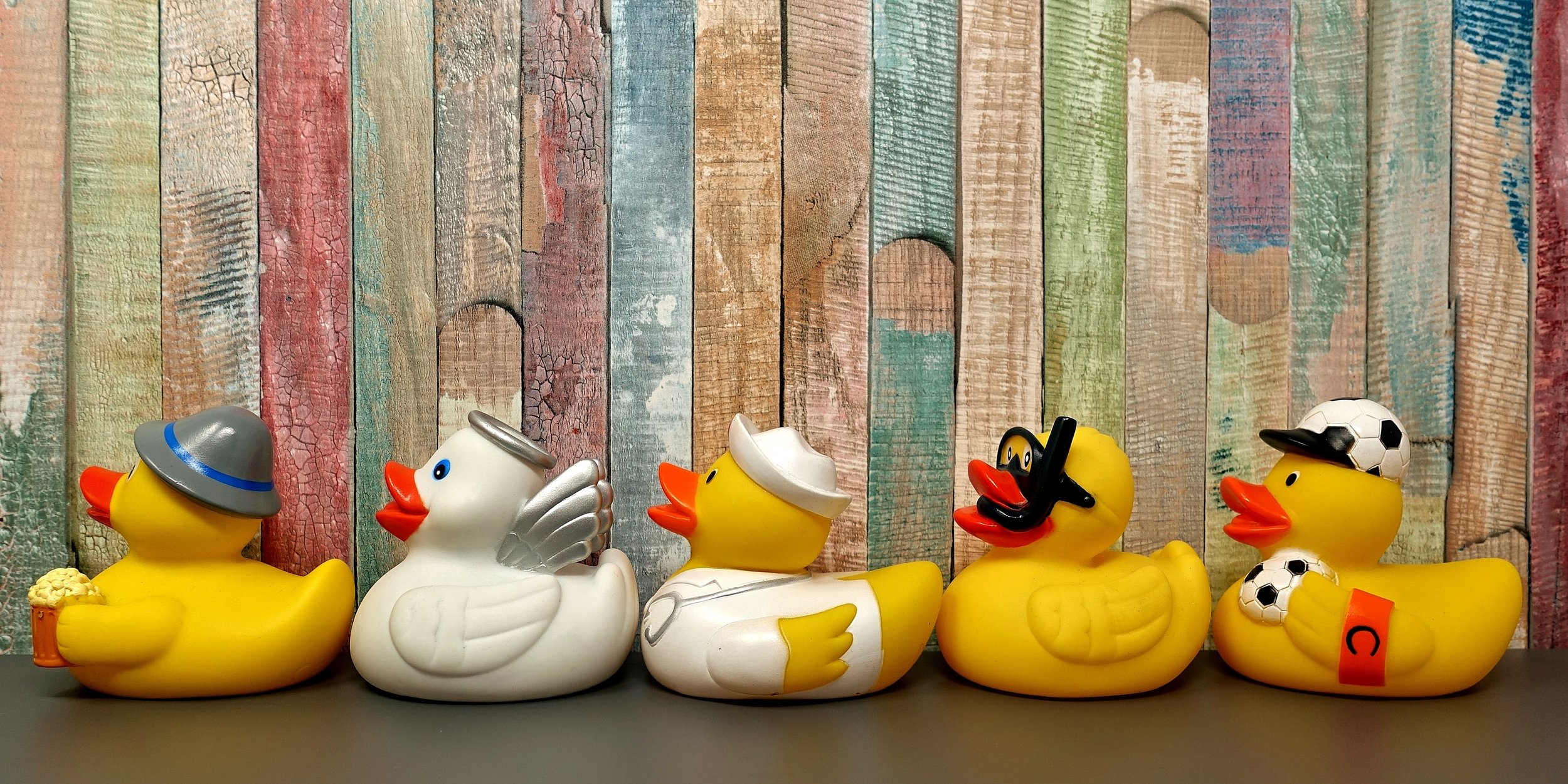 Five yellow rubber ducks with various themed accessories and costumes, lined up on a black surface against a colorful wooden plank background.