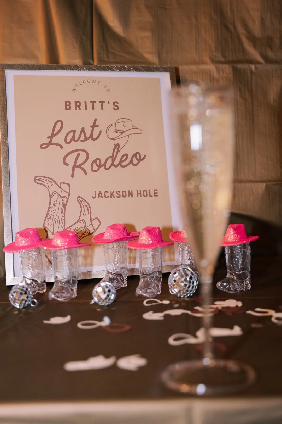 Party table with a sign reading 'Britt's Last Rodeo' at Jackson Hole, pink miniature cowboy hats, small disco ball ornaments, and a blurred champagne glass in the foreground.