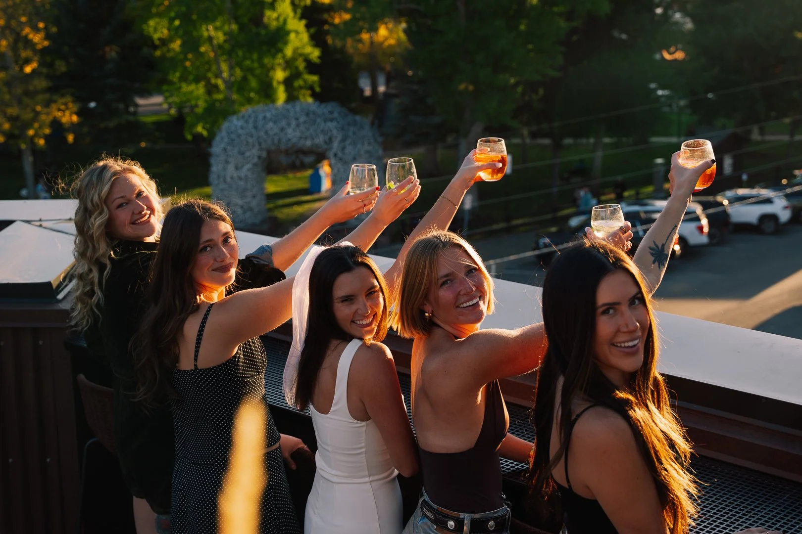 Group of six women celebrating on a rooftop at sunset, raising glasses of drinks in a toast with a city parking lot and trees in the background.