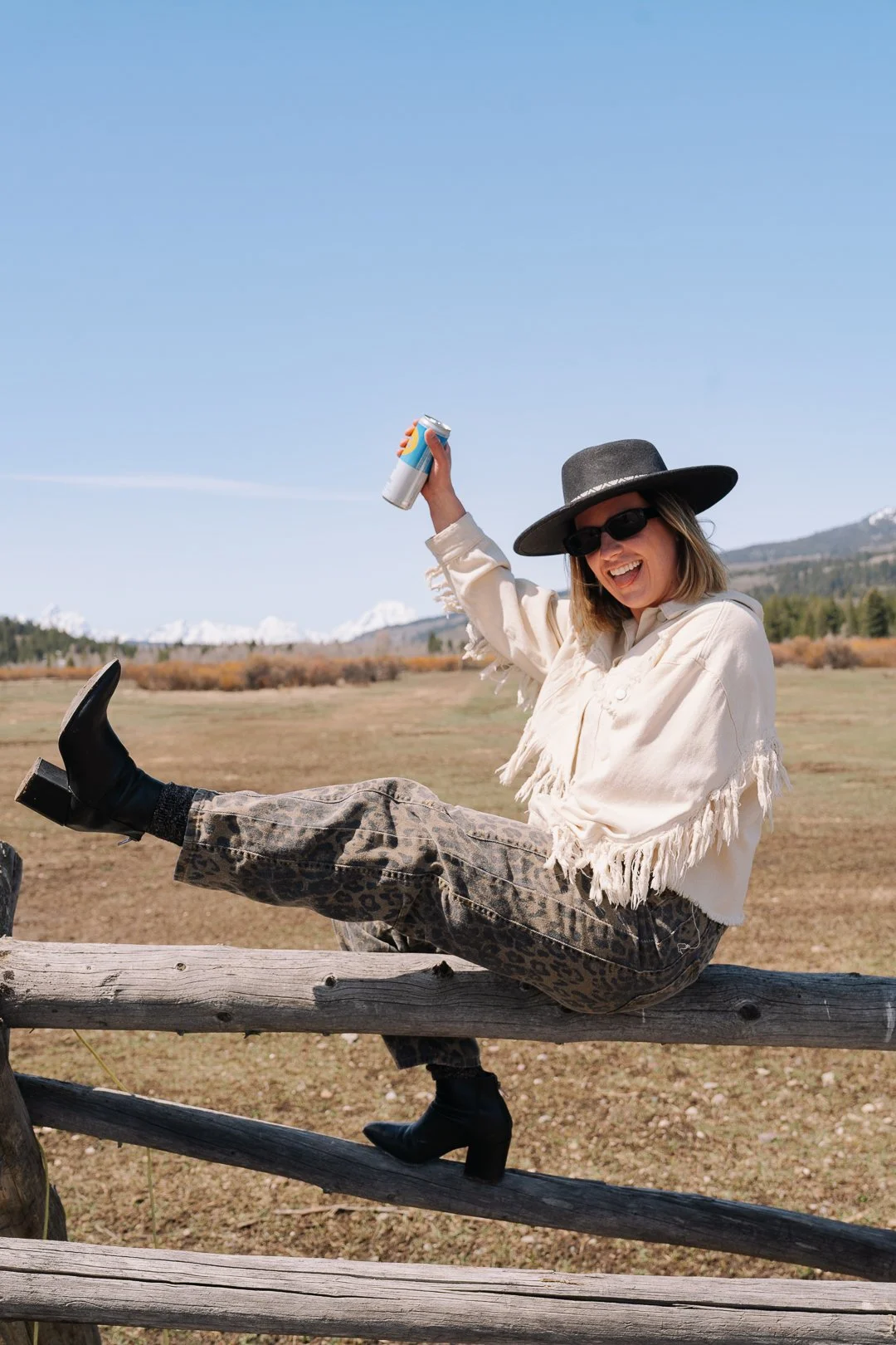A woman sitting on a wooden fence in an open field, holding a drink can in one hand, wearing a wide-brimmed hat, sunglasses, a cream-colored fringed jacket, leopard print pants, and black heeled boots, smiling and enjoying a sunny day.
