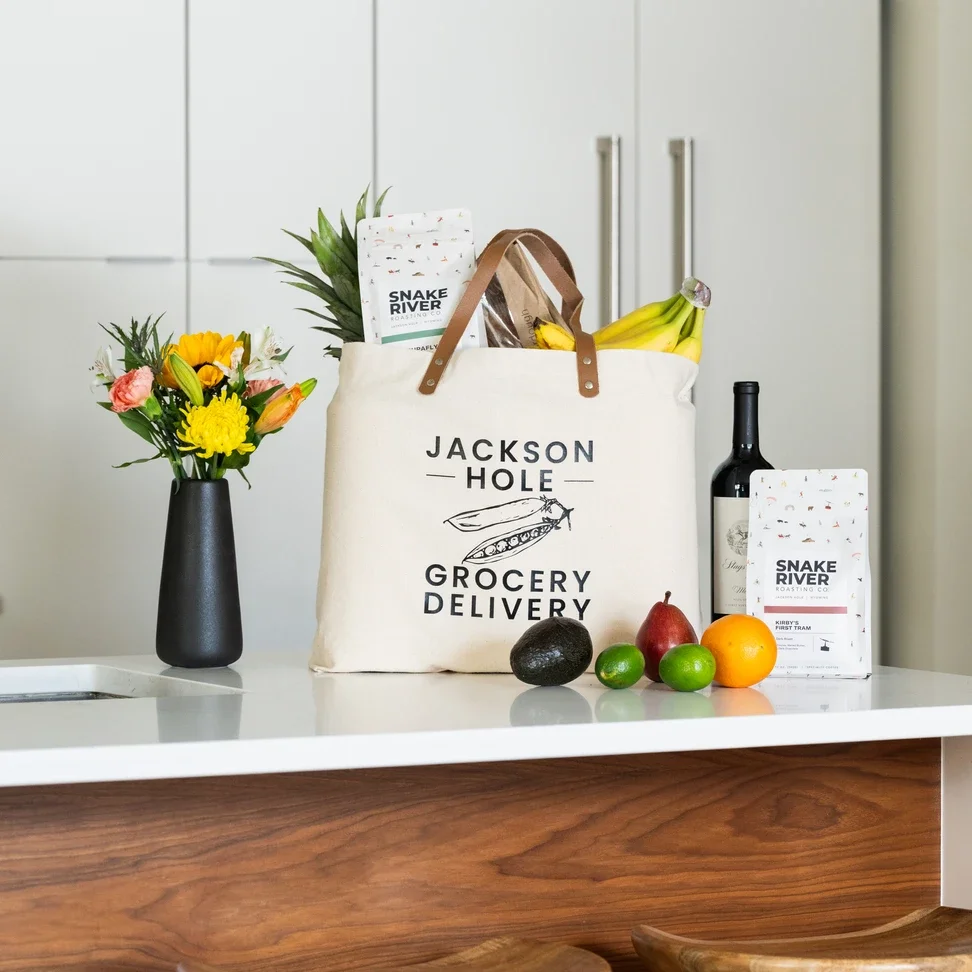 Grocery items on a kitchen white countertop, including a floral arrangement, a tote bag labeled "Jackson Hole Grocery Delivery" filled with produce and packaged goods, fresh fruit such as bananas, avocados, a pear, and an orange, a bottle of wine, and a bag of coffee labeled "Snake River Roasting Co."