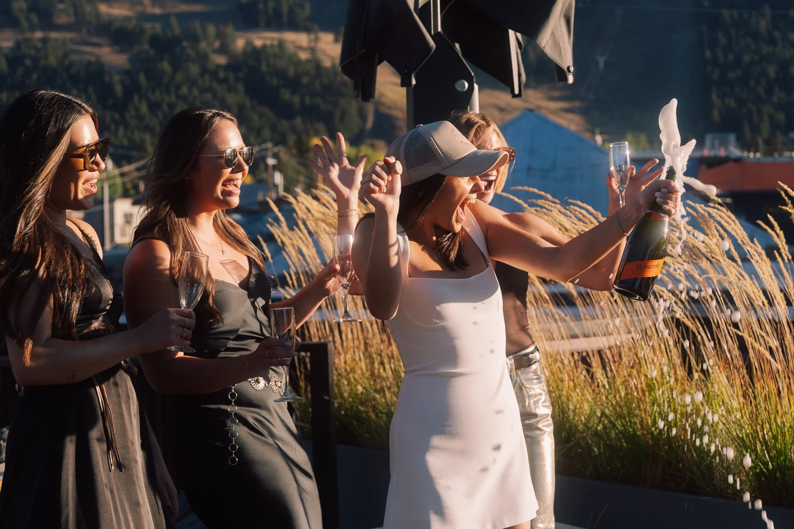 Women celebrating outdoors with champagne, some wearing sunglasses, with one woman opening a champagne bottle and everyone holding glasses.