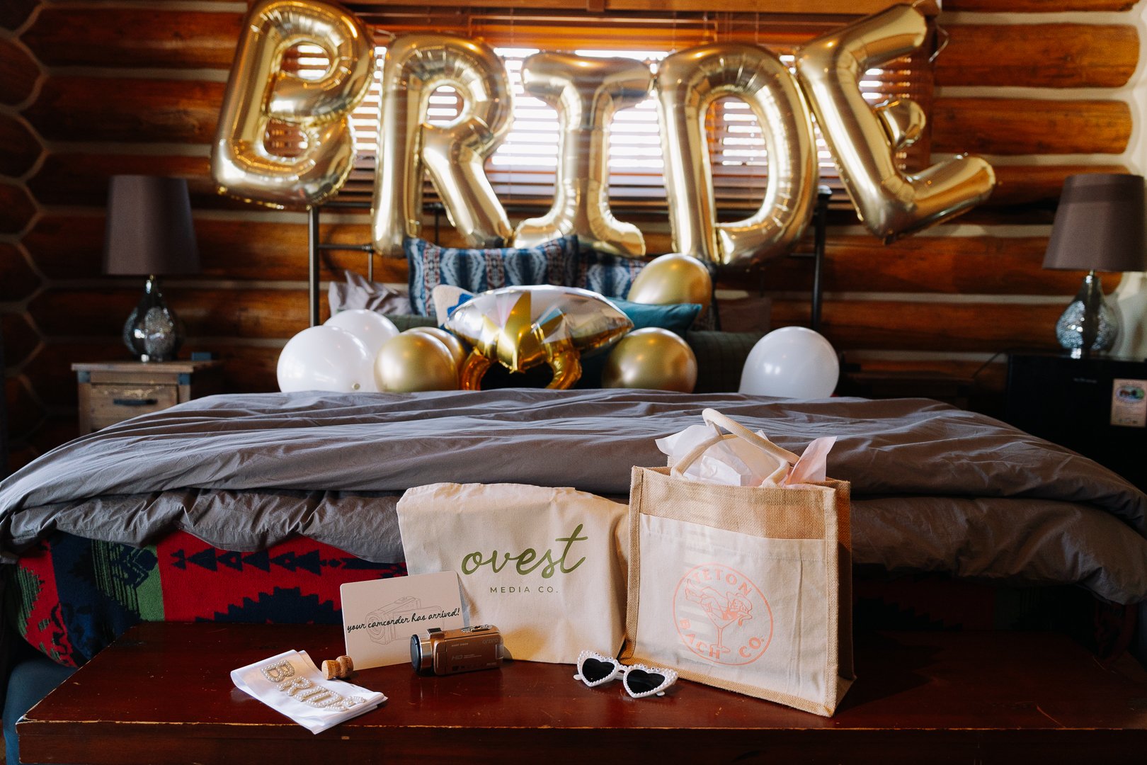 A bedroom decorated with gold balloons spelling out "BRED" and additional balloons, some in shapes and others in solid colors, arranged over the bed. The bed is made with gray bedding, and there are pillows, a blanket, and decorative elements, including sunglasses and tote bags on a wooden table in front of the bed. The room has wooden walls and side tables with lamps.