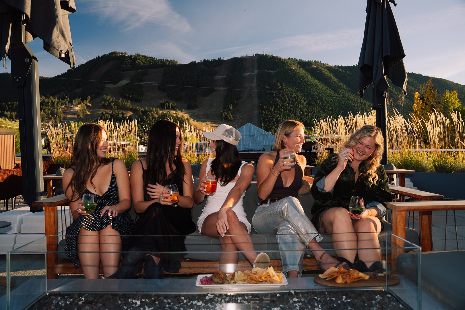 Five women sitting on a couch outdoors, smiling, holding drinks, and talking, with a mountain landscape and a blue sky in the background.