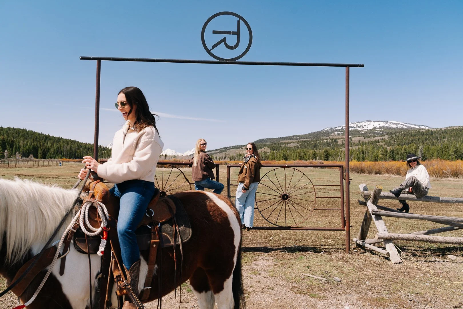 Four women enjoying a sunny day outdoors at a rural ranch with mountains and snow in the background. One woman is riding a horse, wearing sunglasses and smiling. Two women are standing near a gate, facing the camera, and one woman is sitting on a wooden fence wearing a black hat.