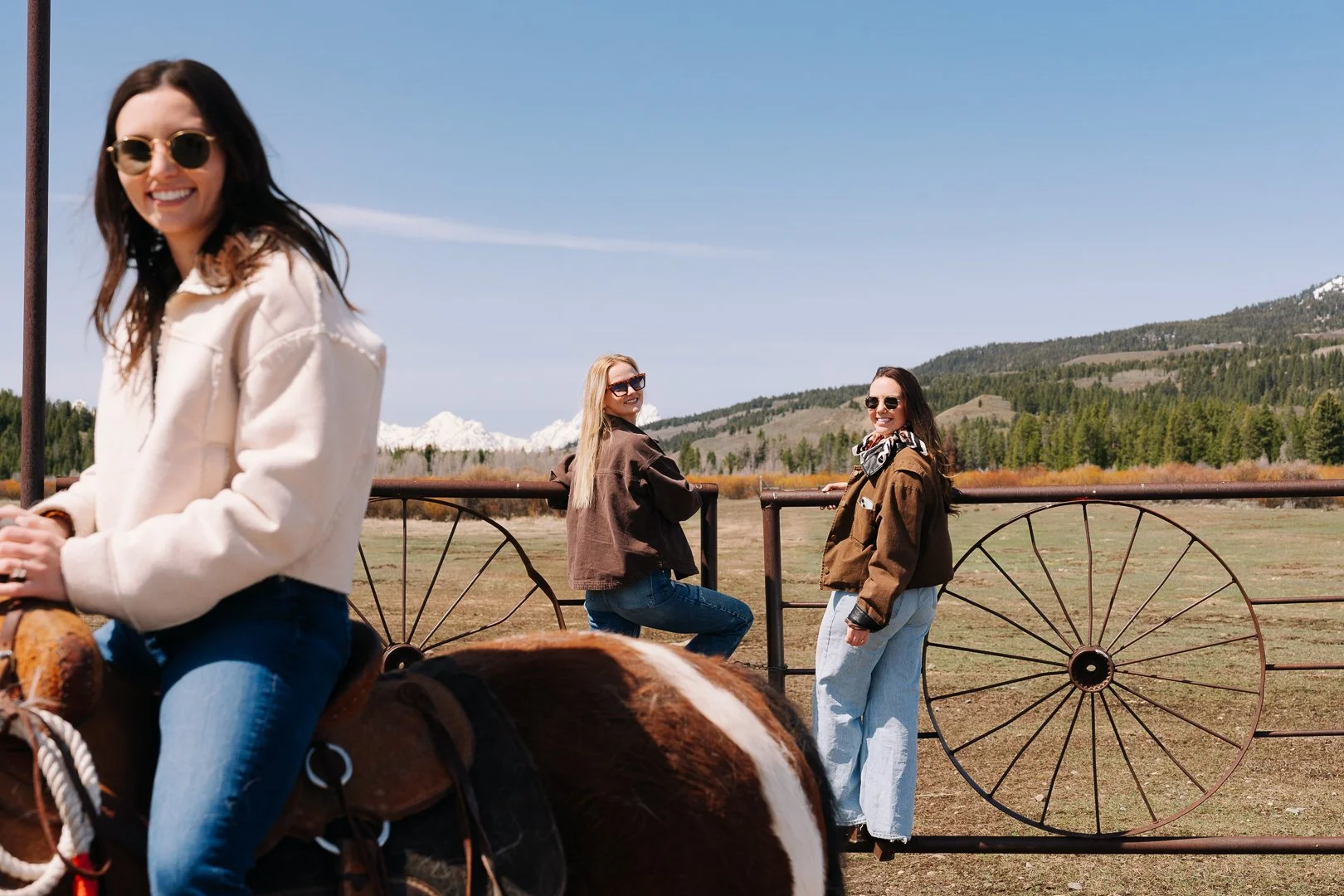 Three women outdoors in a rural setting with mountains in the background. One woman is riding a horse, and two women are standing by a metal fence, all smiling and wearing sunglasses.