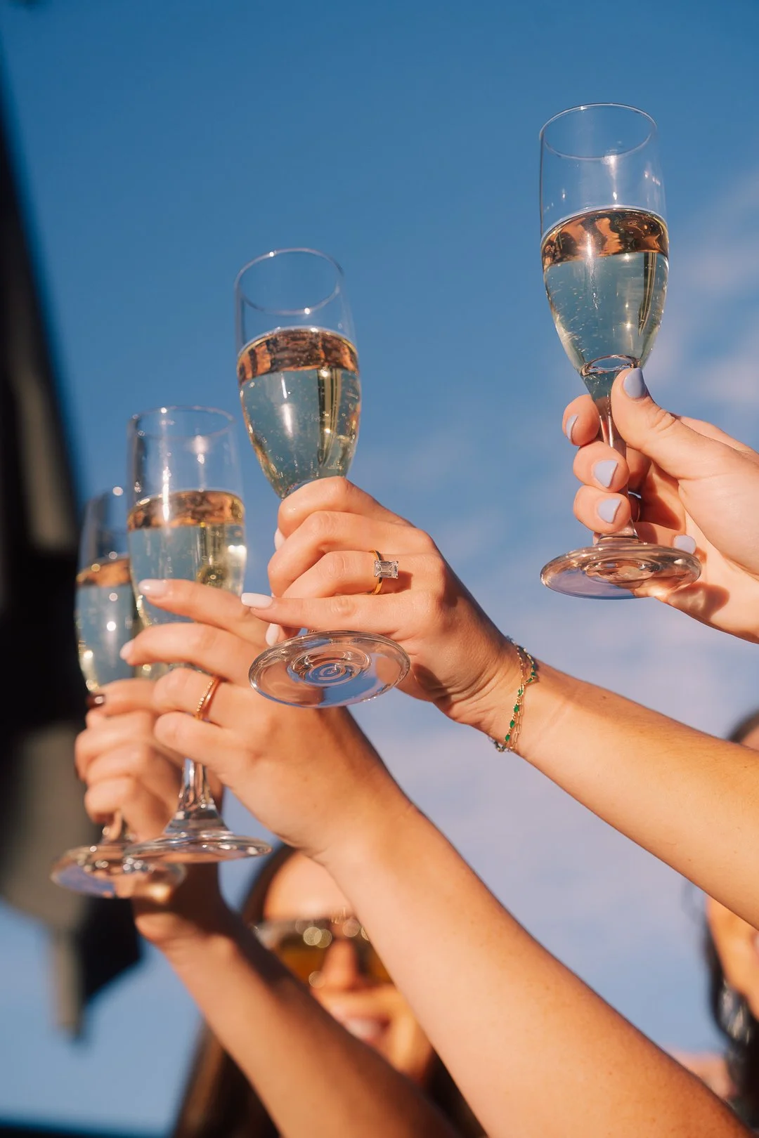 Multiple people holding champagne flutes filled with champagne in a toast outdoors against a blue sky.