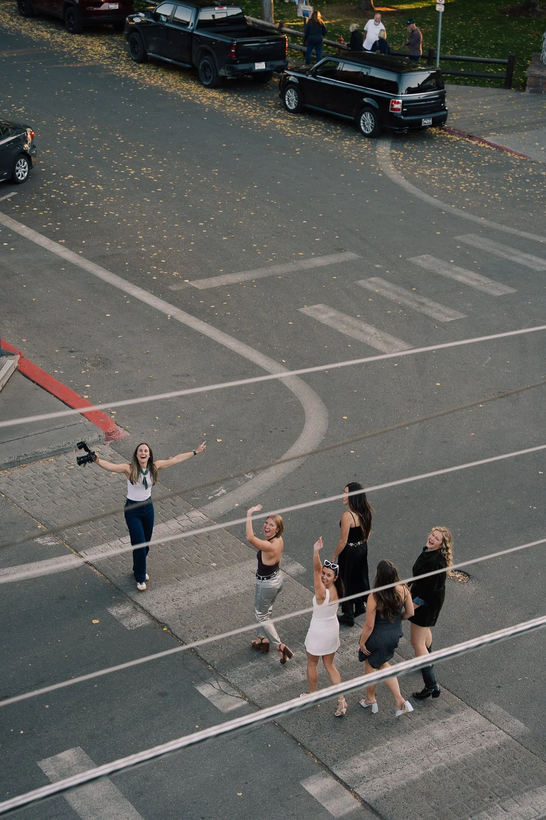 Six women standing on a city sidewalk, smiling and waving at the camera, with one woman holding a video camera. They are dressed in casual and semi-formal attire, celebrating or socializing outside on a fall day with leaves scattered on the ground.