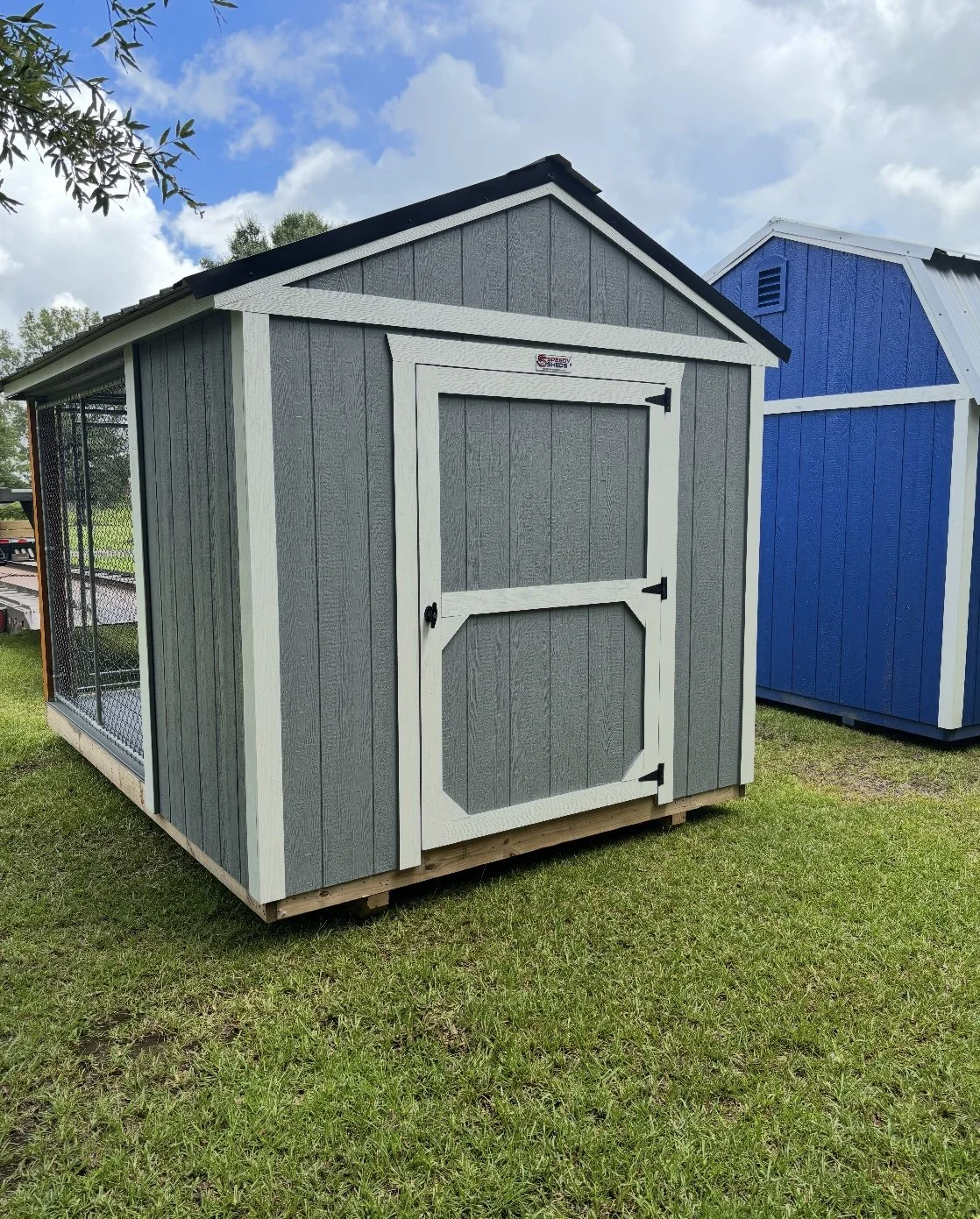Gray custom shed with a white-trimmed door and attached kennel run, displayed on a grassy lot.