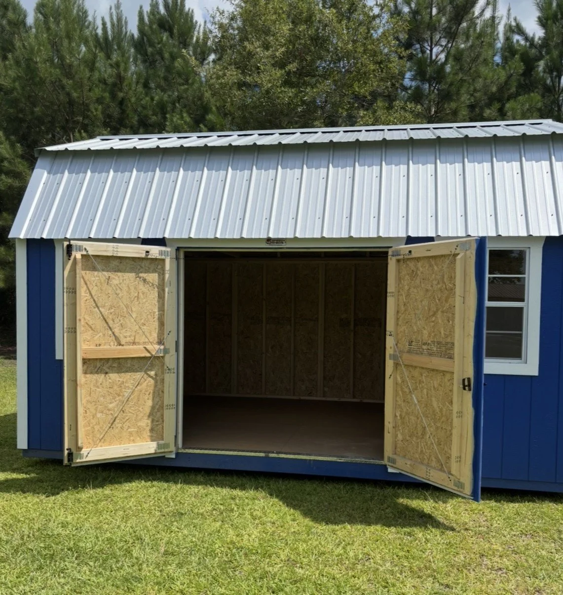 Blue barn-style storage shed with double doors open, showing an unfinished wooden interior and front-facing windows.