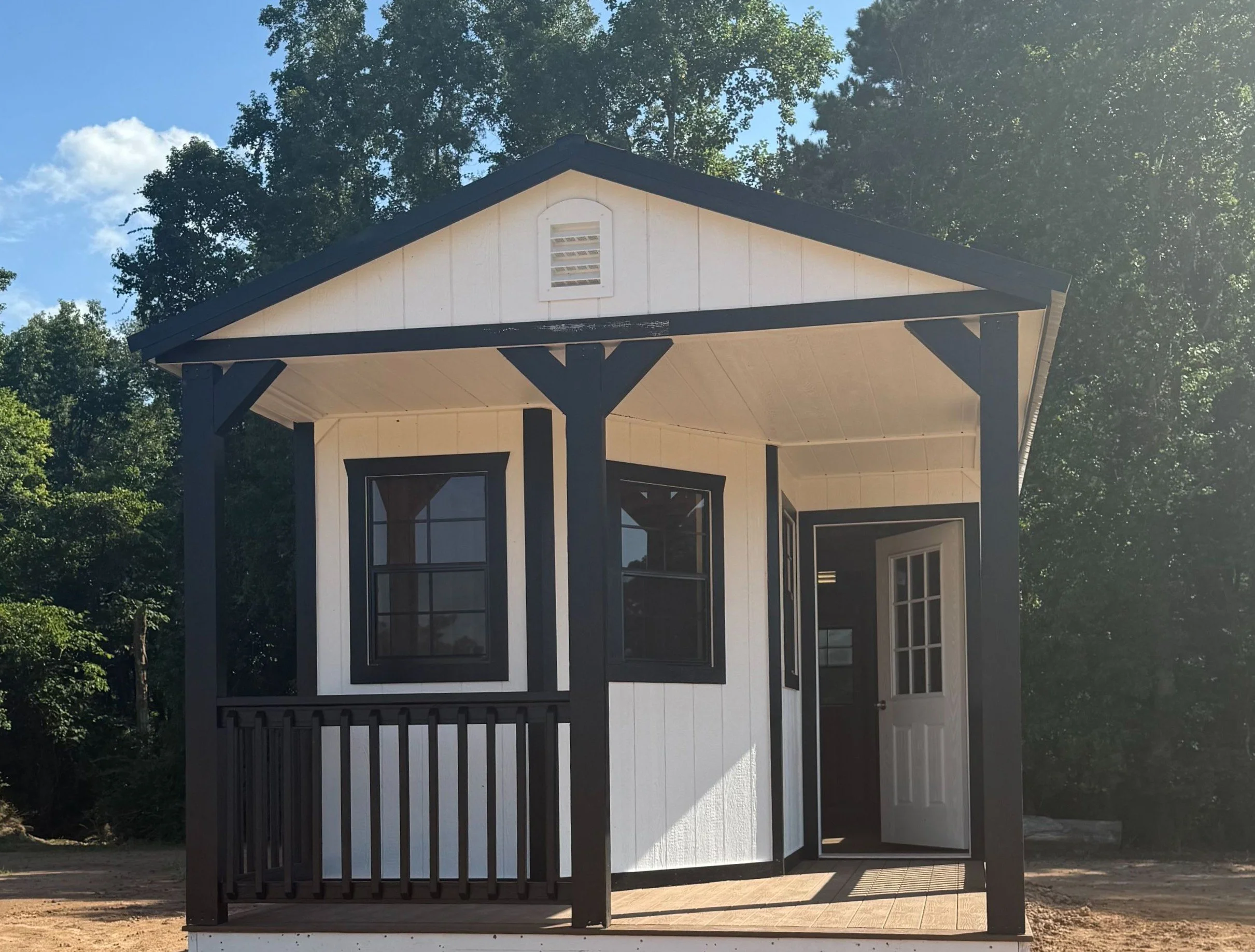 Modern white portable cabin with black trim, featuring a front porch, black railing, and open door under a gable roof.
