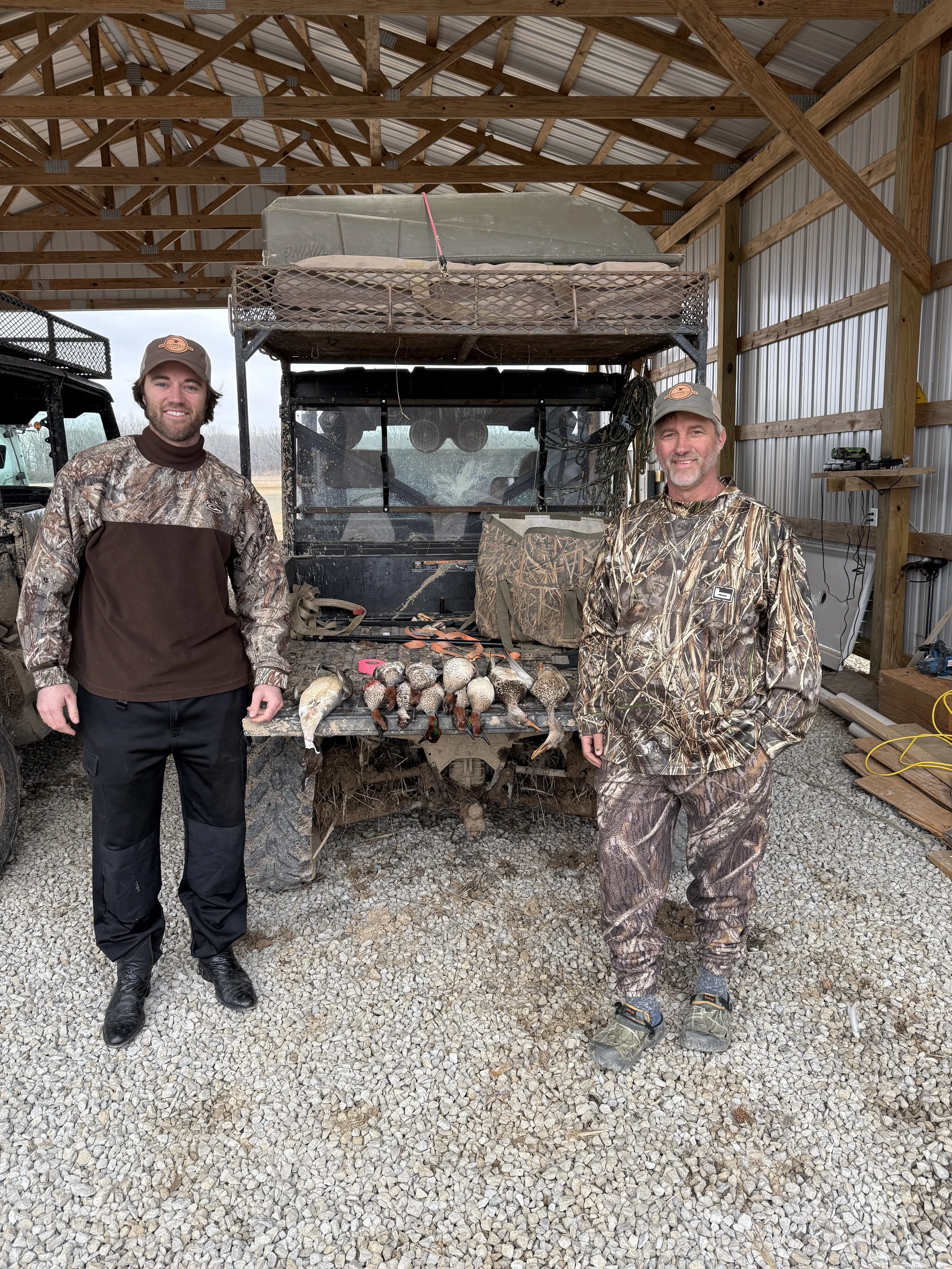 Two men standing in front of a utility vehicle with a rack of ducks and a dead fish, inside a wooden shed with a gravel floor.