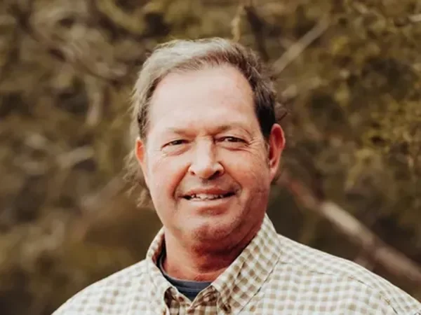 A man smiling outdoors, wearing a checkered button-up shirt with a background of trees and foliage.