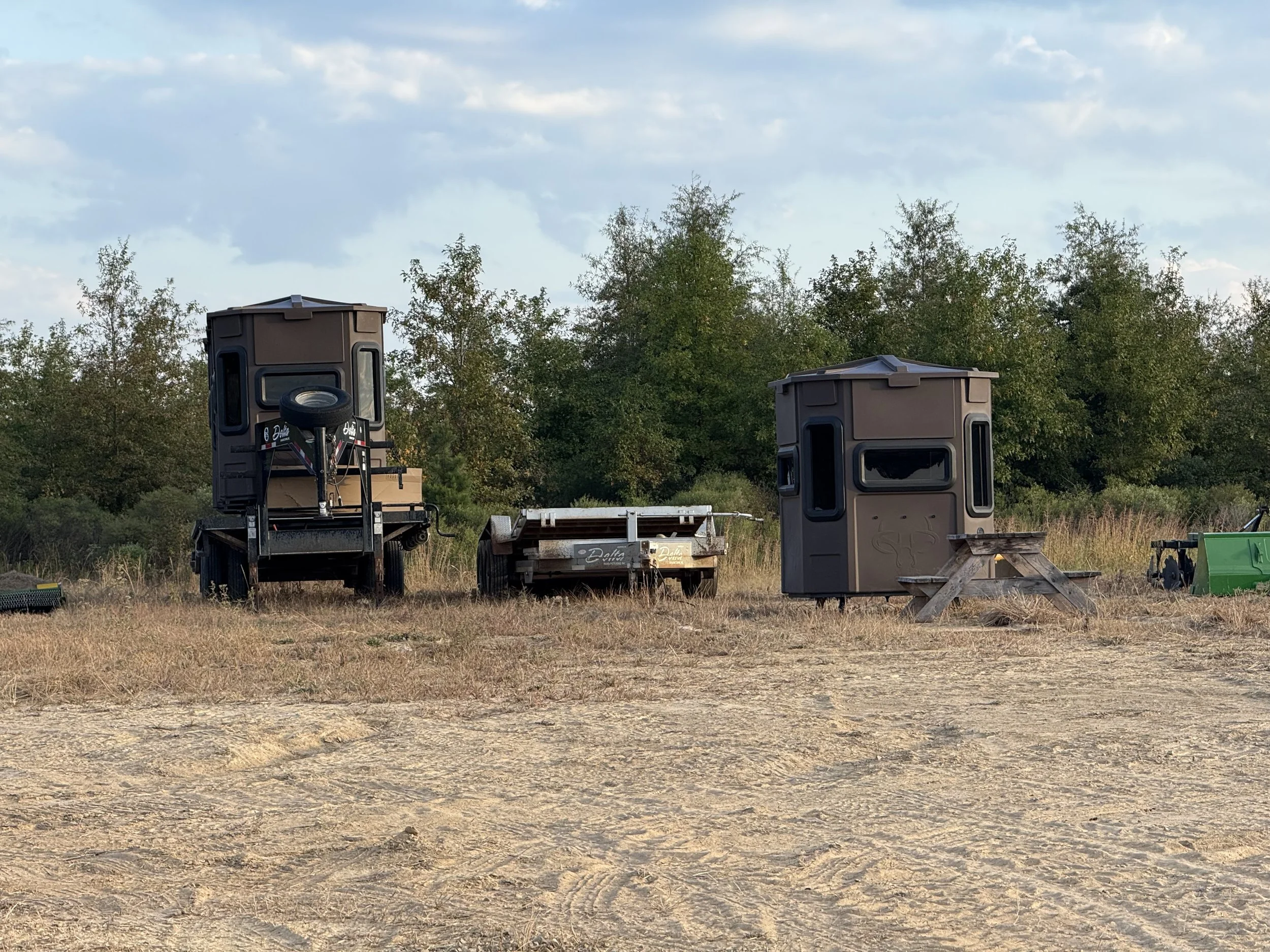 Various hunting blinds and equipment set up on a field with trees in the background and a cloudy sky.