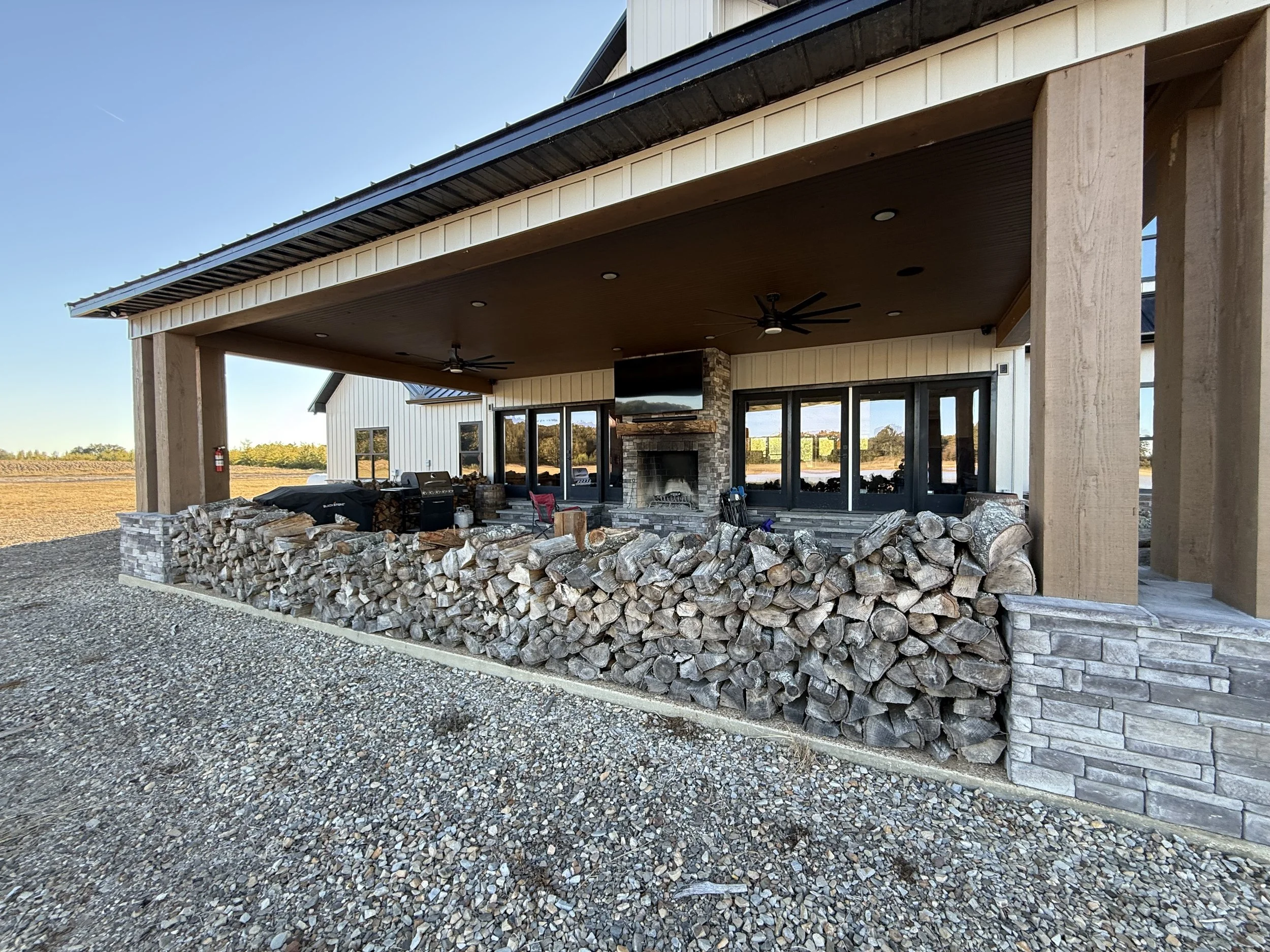 Large outdoor patio area attached to a modern house, featuring a stone and wood firewood storage wall, outdoor fireplace, ceiling fans, and multiple glass doors leading inside. Gravel ground surrounds the patio, with open fields visible in the backgr