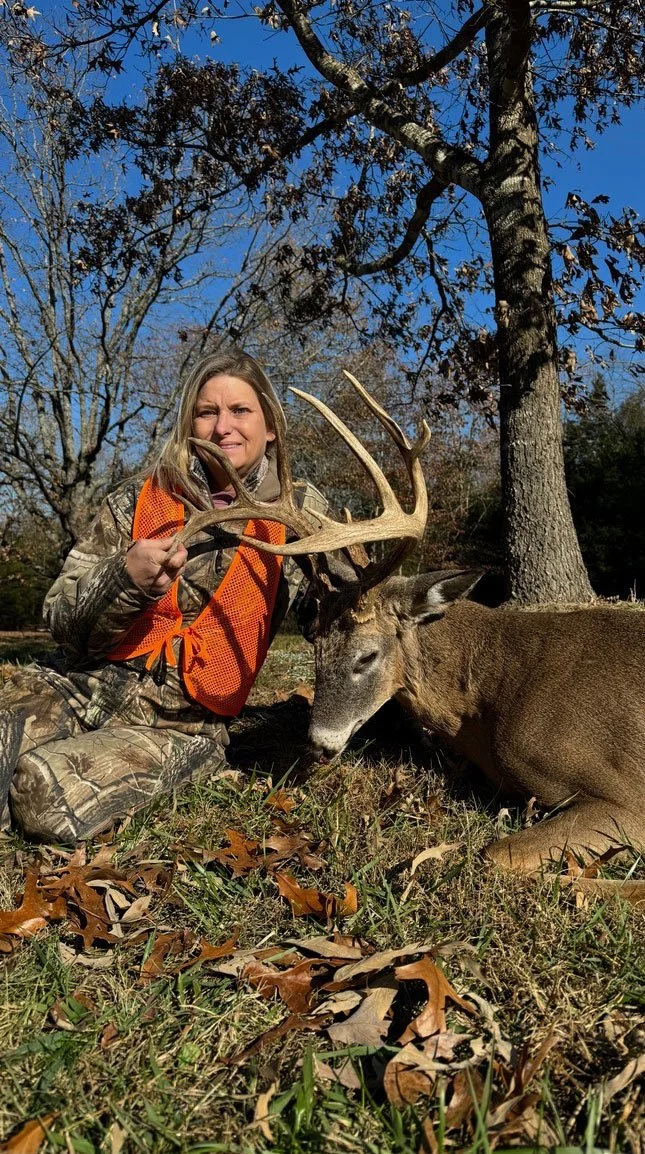 Woman in camouflage clothing and orange safety vest holding antlers of a dead deer, lying on grass with fallen leaves in a wooded area.