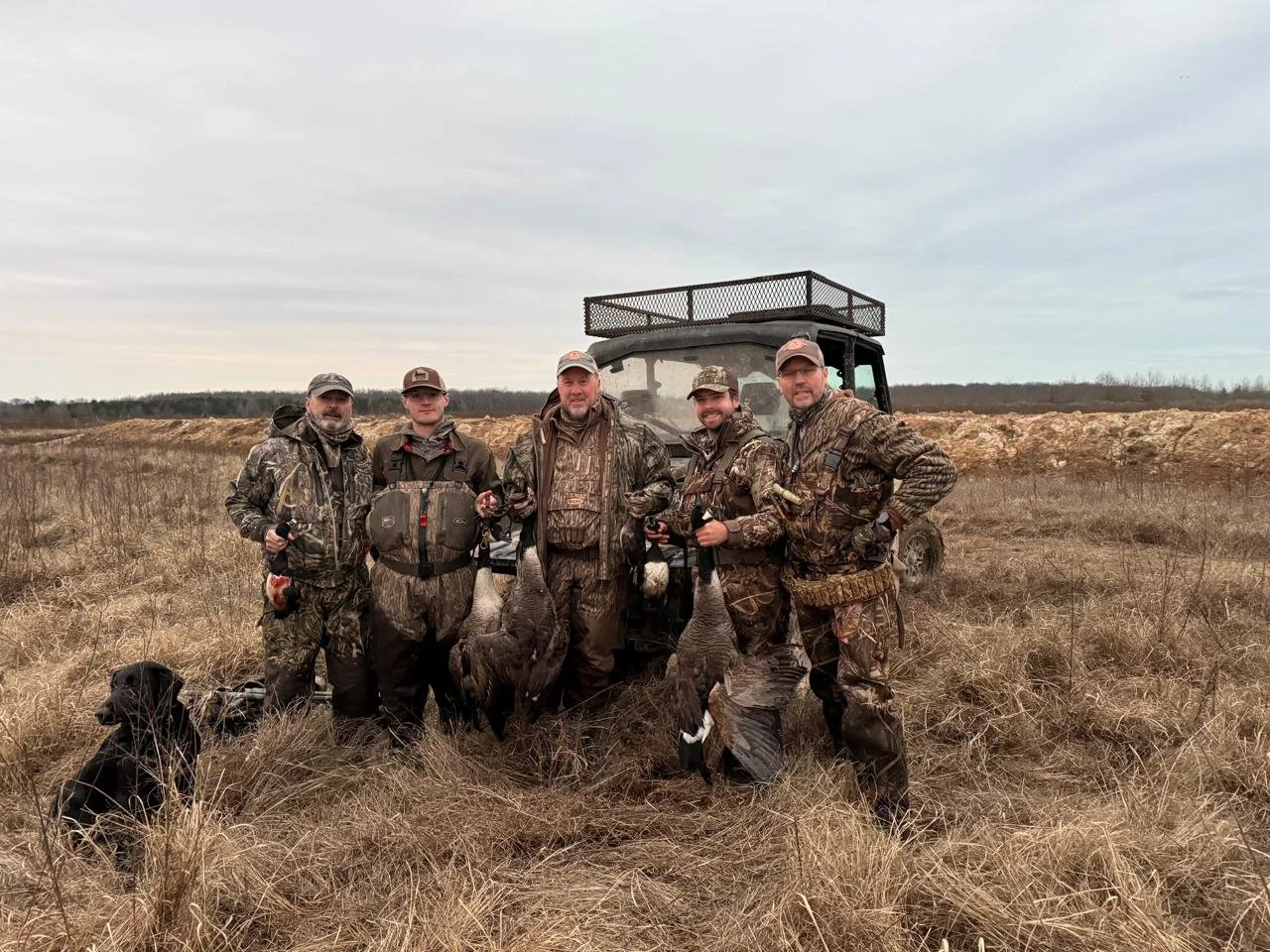 Five men in camouflage gear posing in a grassy field with hunting decoys and a golf cart, holding duck decoys and hunting equipment, alongside a black dog.