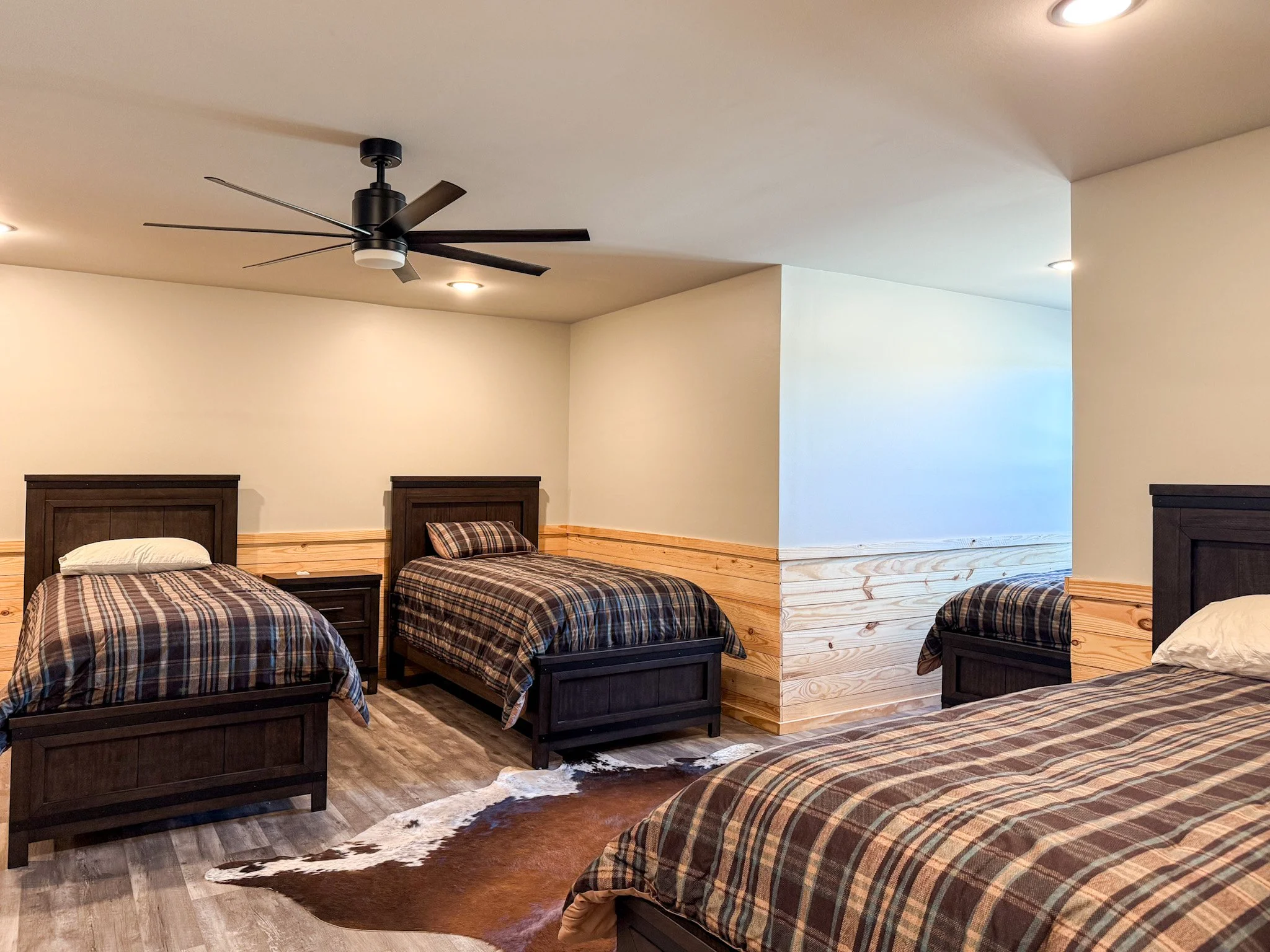 A bedroom with two beds featuring plaid bedding, a window with black mesh blinds, a black cowhide rug, a ceiling fan, and wood paneling on the walls.