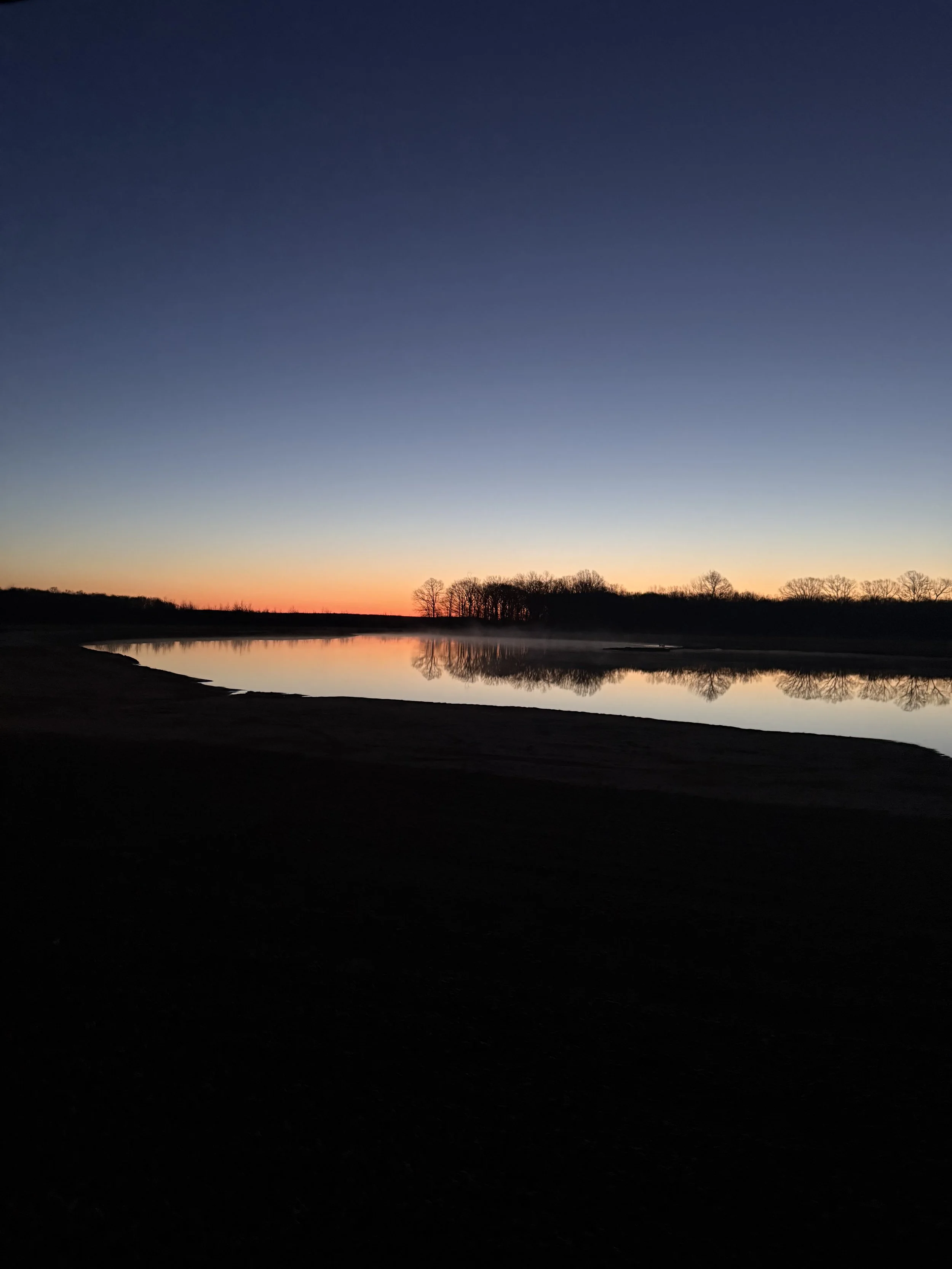 Sunset over a calm river with reflections of trees and a dark shoreline in the foreground.