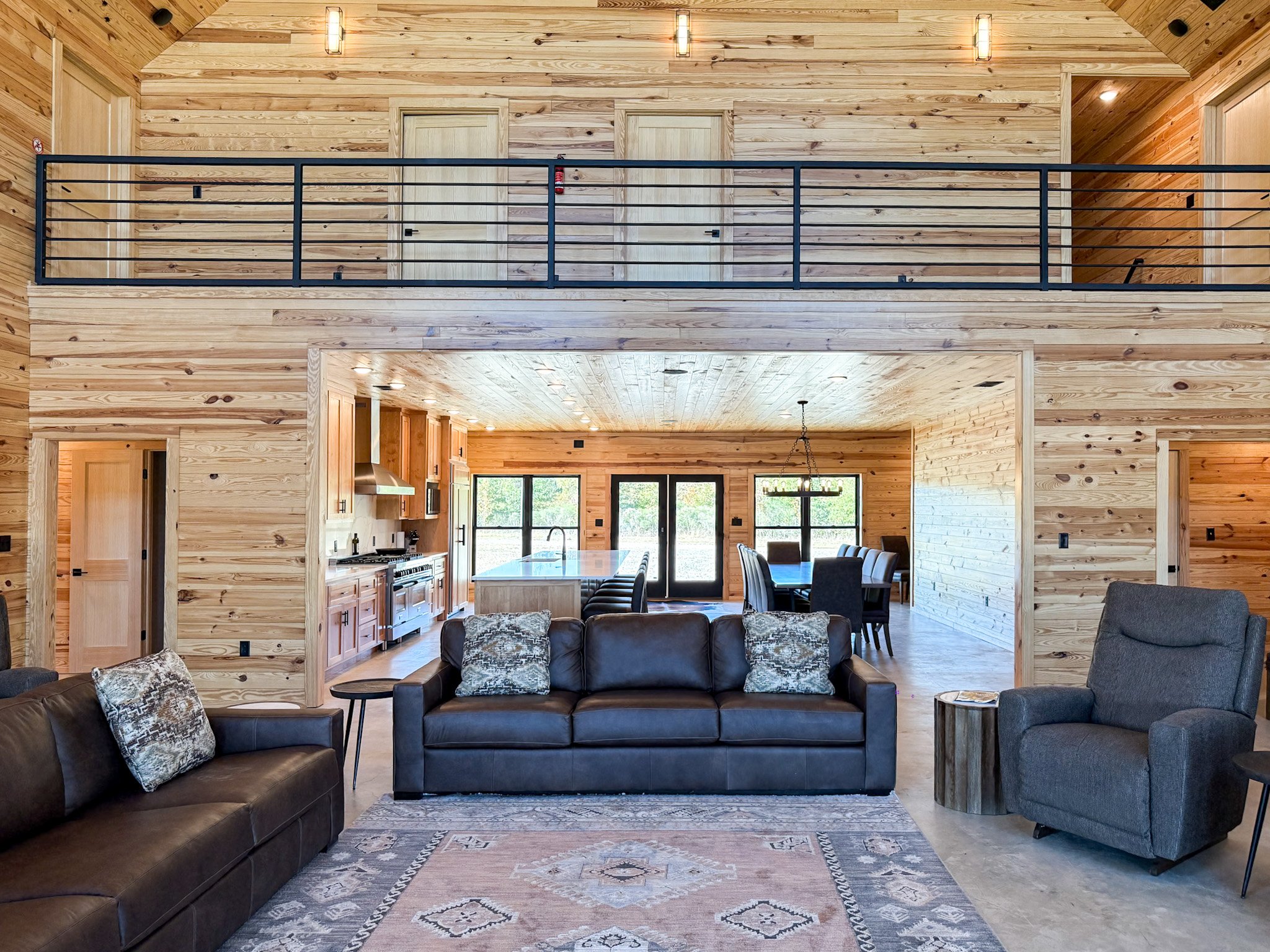 A spacious living room with high wooden walls and ceiling, featuring a large stone fireplace, multiple windows, two antler chandeliers, and various sofas and chairs arranged for seating.