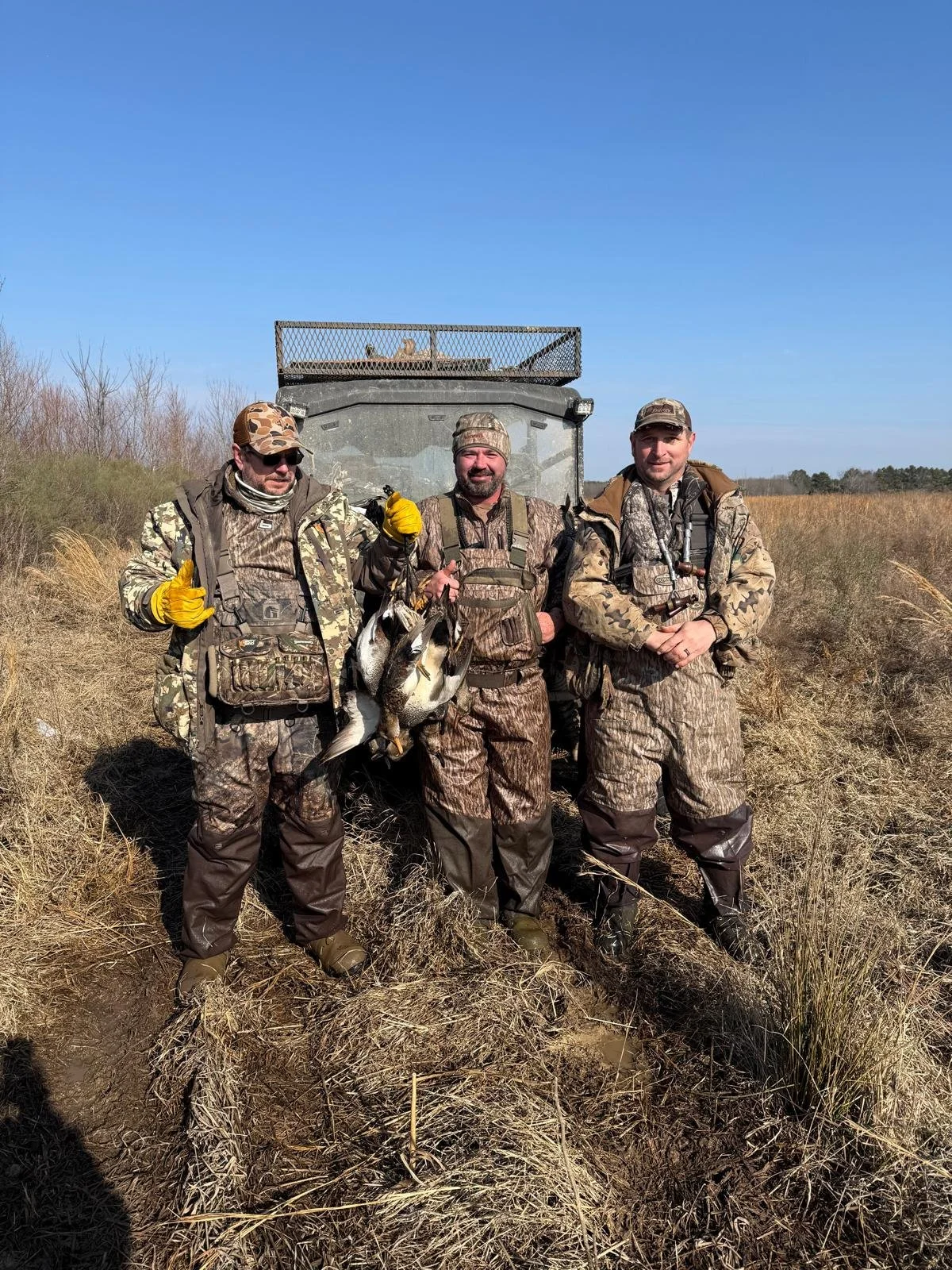 Three men in camouflage hunting gear standing in a grassy field, holding ducks they hunted, with a utility vehicle in the background under a blue sky.