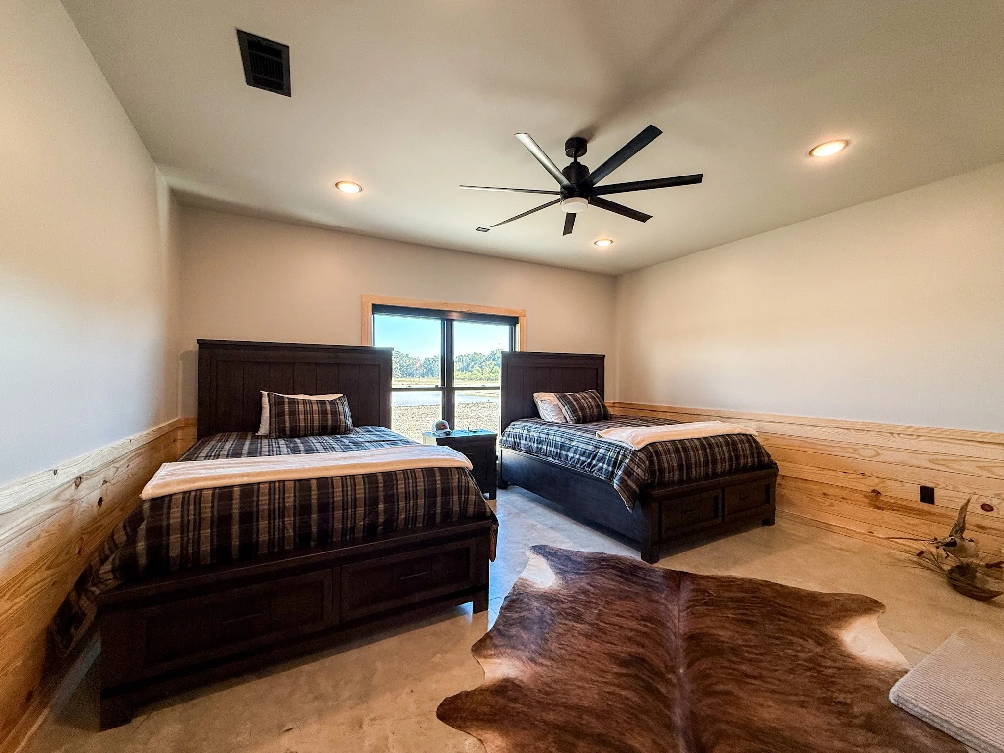 A bedroom with a dark wooden bed frame and plaid bedding, wooden paneling on the lower half of the walls, and a beige carpet.