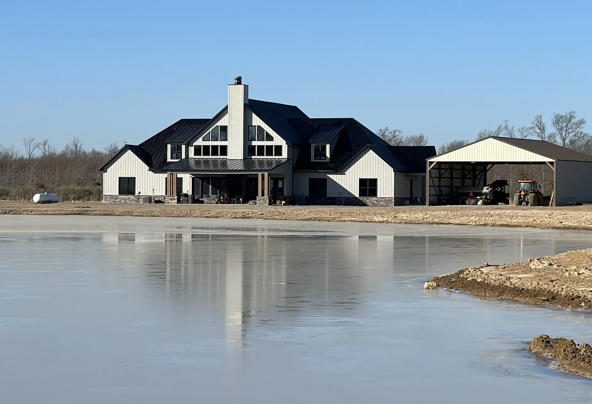 A modern house under construction with a black metal roof, metal siding, and large windows, situated next to a body of water with a frozen surface, and a covered area with construction equipment and a tractor nearby.
