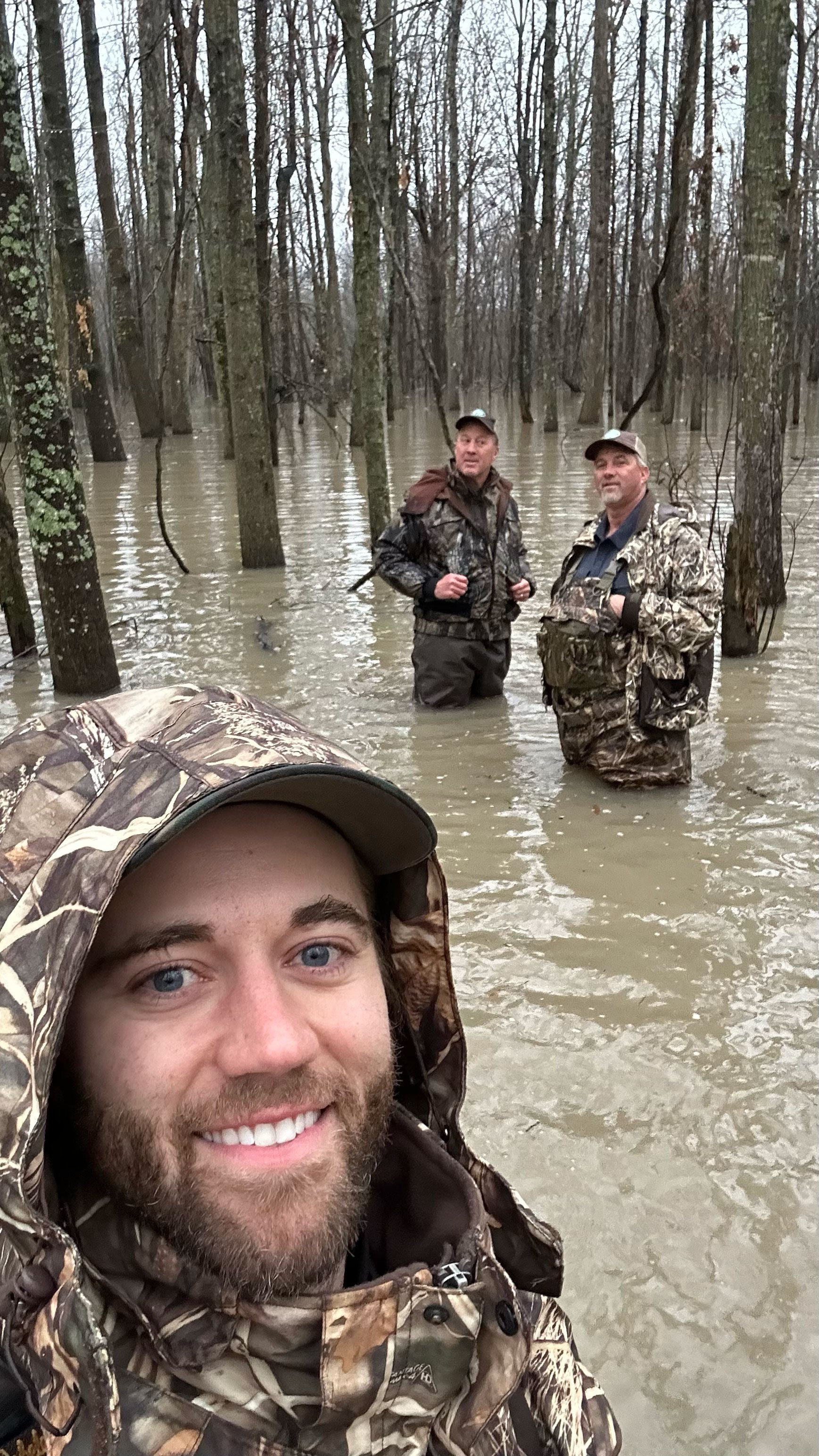 Three men in camouflage and outdoor gear stand in a flooded forest with leafless trees, taking a selfie with a man in the foreground smiling.