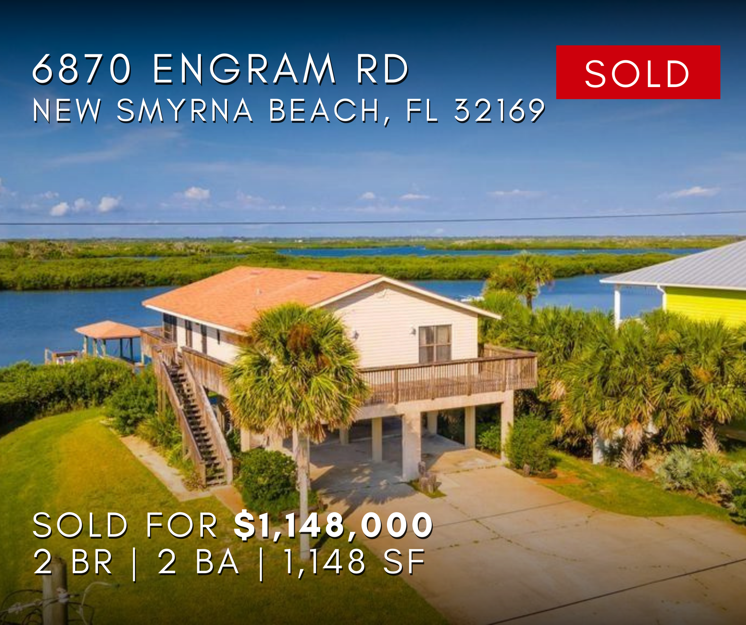 New Smyrna Beach waterfront home property, 6870 Engram road nsb, Florida A house with a white exterior, red roof, and wooden deck, elevated on stilts, surrounded by palm trees, near water, with a driveway in front. Beach Life