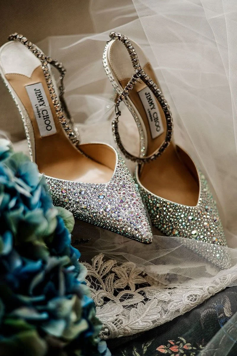 Elegante silberne High Heels mit funkelnden Steinen, auf einem weißen Tüllstoff mit Spitze liegend, für eine Hochzeit geeignet.