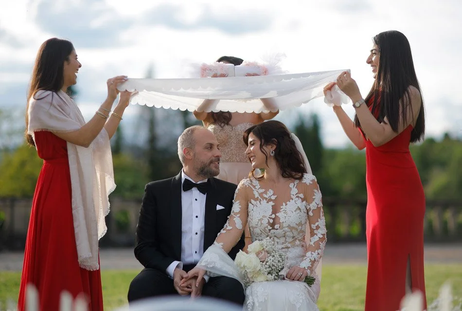 Hochzeit im Freien mit einem Brautpaar, das auf einer Bank sitzt, umgeben von Freundinnen, die einen weißen Schleier oder Tuch halten.