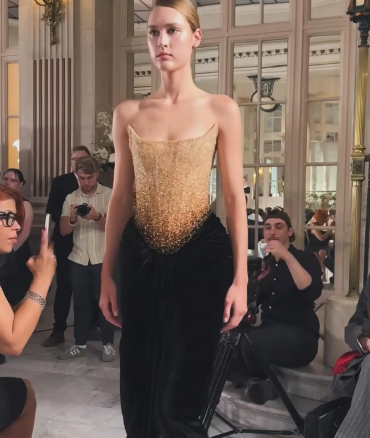 Model walking on runway wearing a strapless, gold and black dress during a fashion show in an elegant venue with audience members taking photos