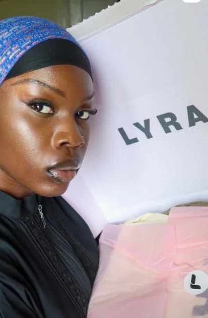A woman wearing a blue headscarf and black jacket taking a selfie next to a white box with the word 'LYRA' on it.