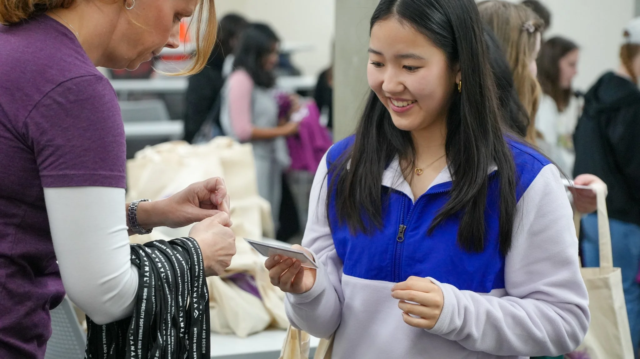 A young woman in a blue and white jacket holding a card, smiling at another person at an indoor event with multiple people in the background.