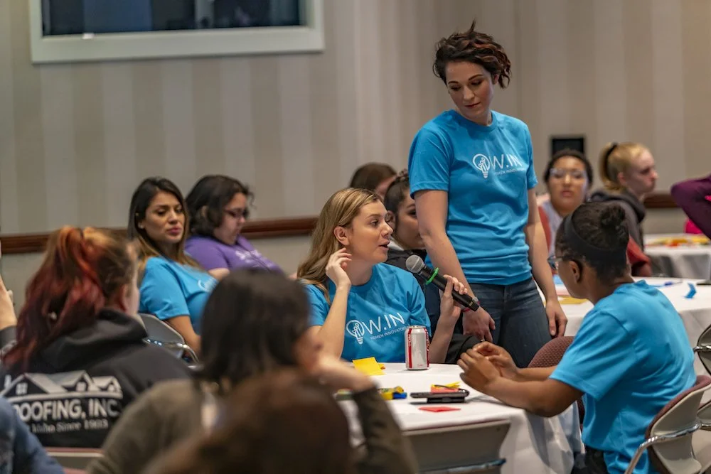 Group of women in a conference room, with a woman holding a microphone and speaking to another woman, while others watch.