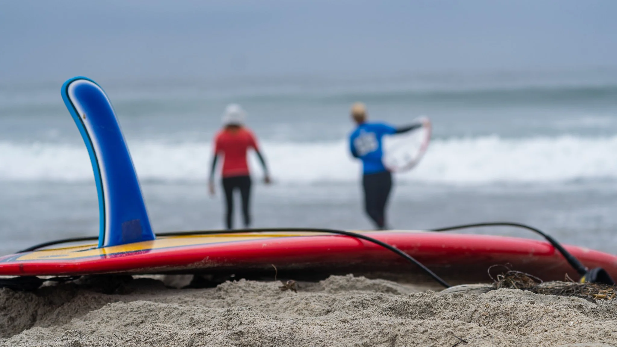 Surfboard and paddle on the sand with two surfers in wetsuits and hats in the background at the beach.