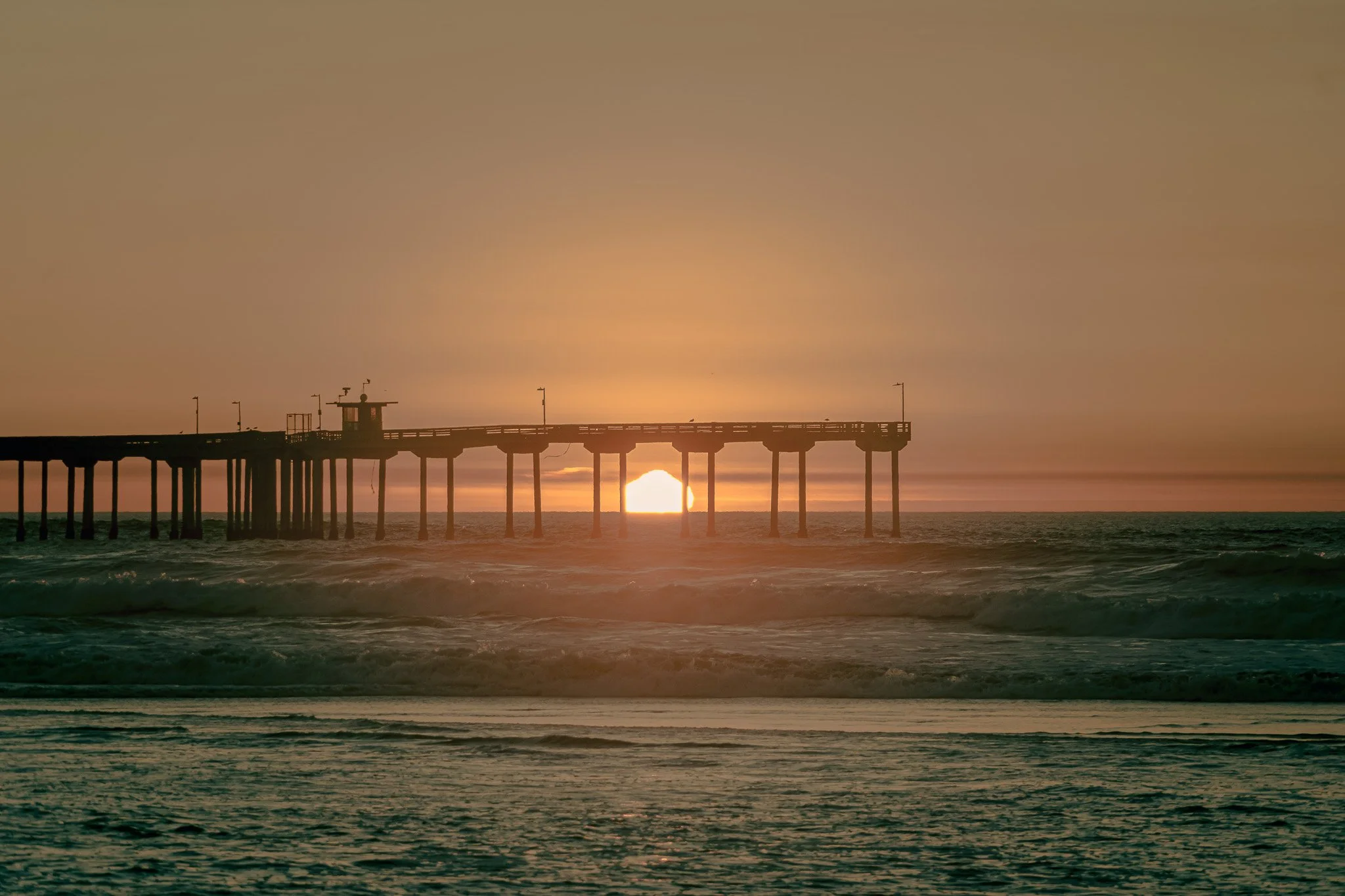 Sunset over a pier extending into the ocean with waves in the foreground.