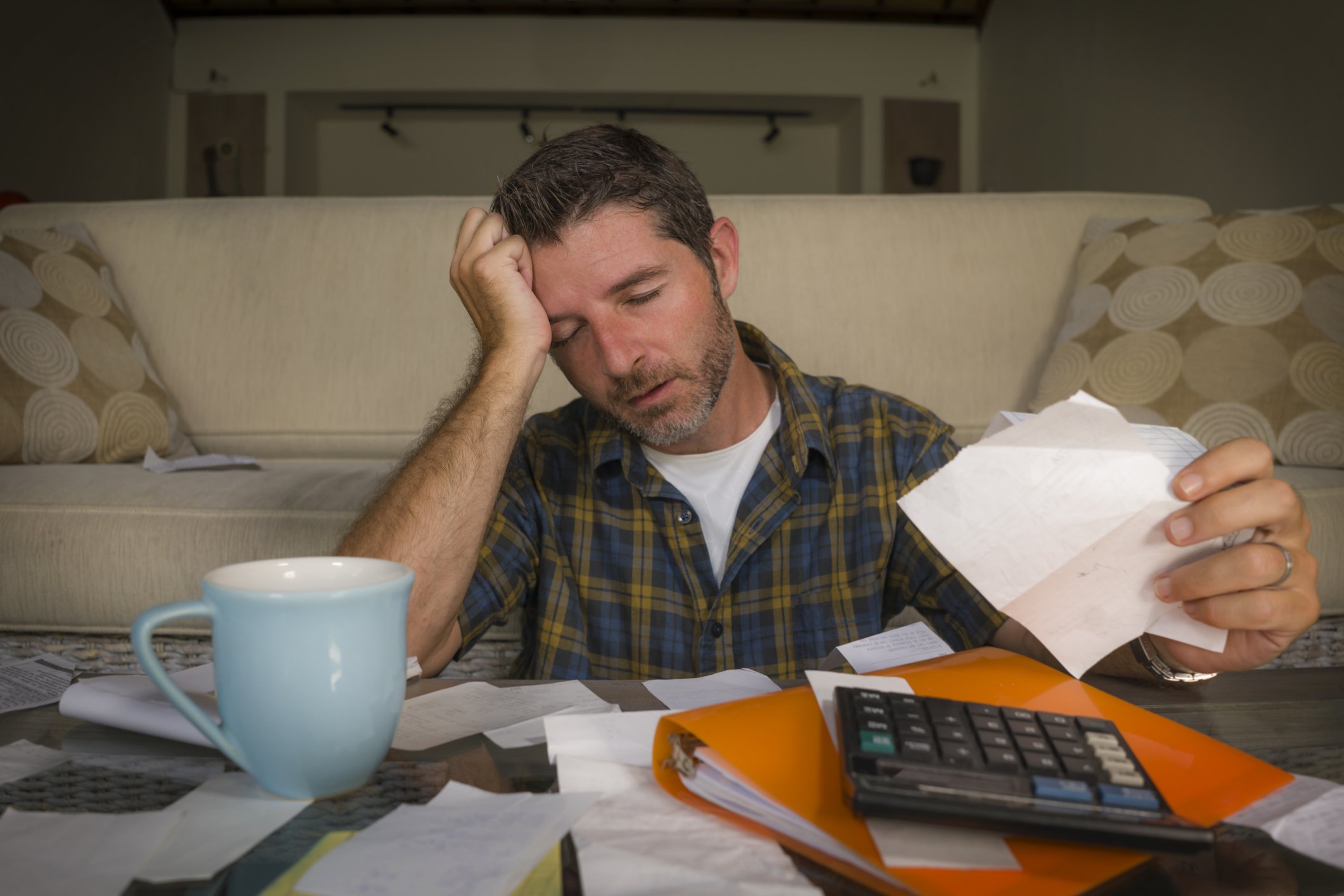 Man sitting at a cluttered table, appearing stressed or overwhelmed while reading papers, with a mug, calculator, and scattered documents around him in a living room