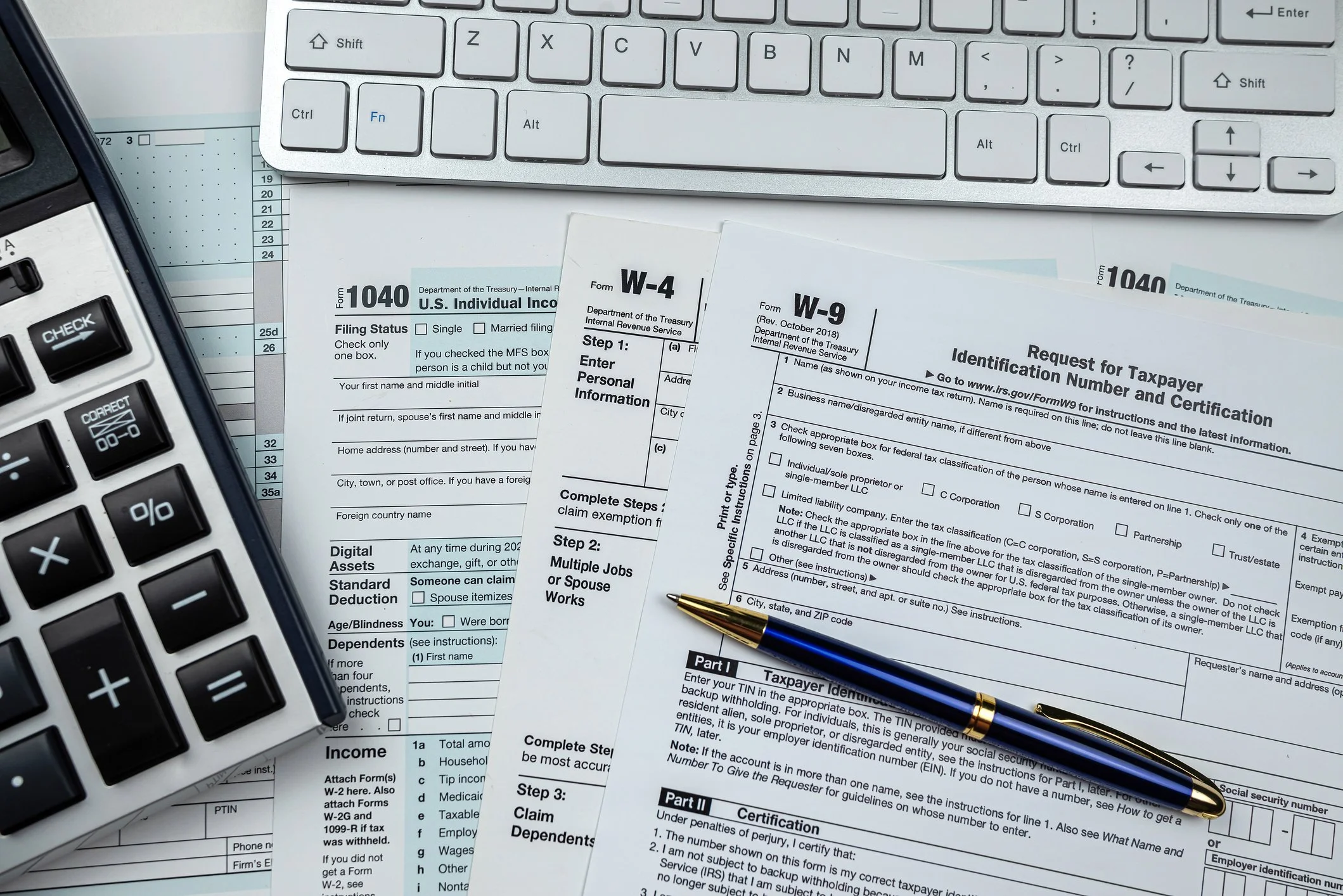 A computer keyboard, tax forms including W-4, W-9, and 1040 on a desk with a pen and a calculator.