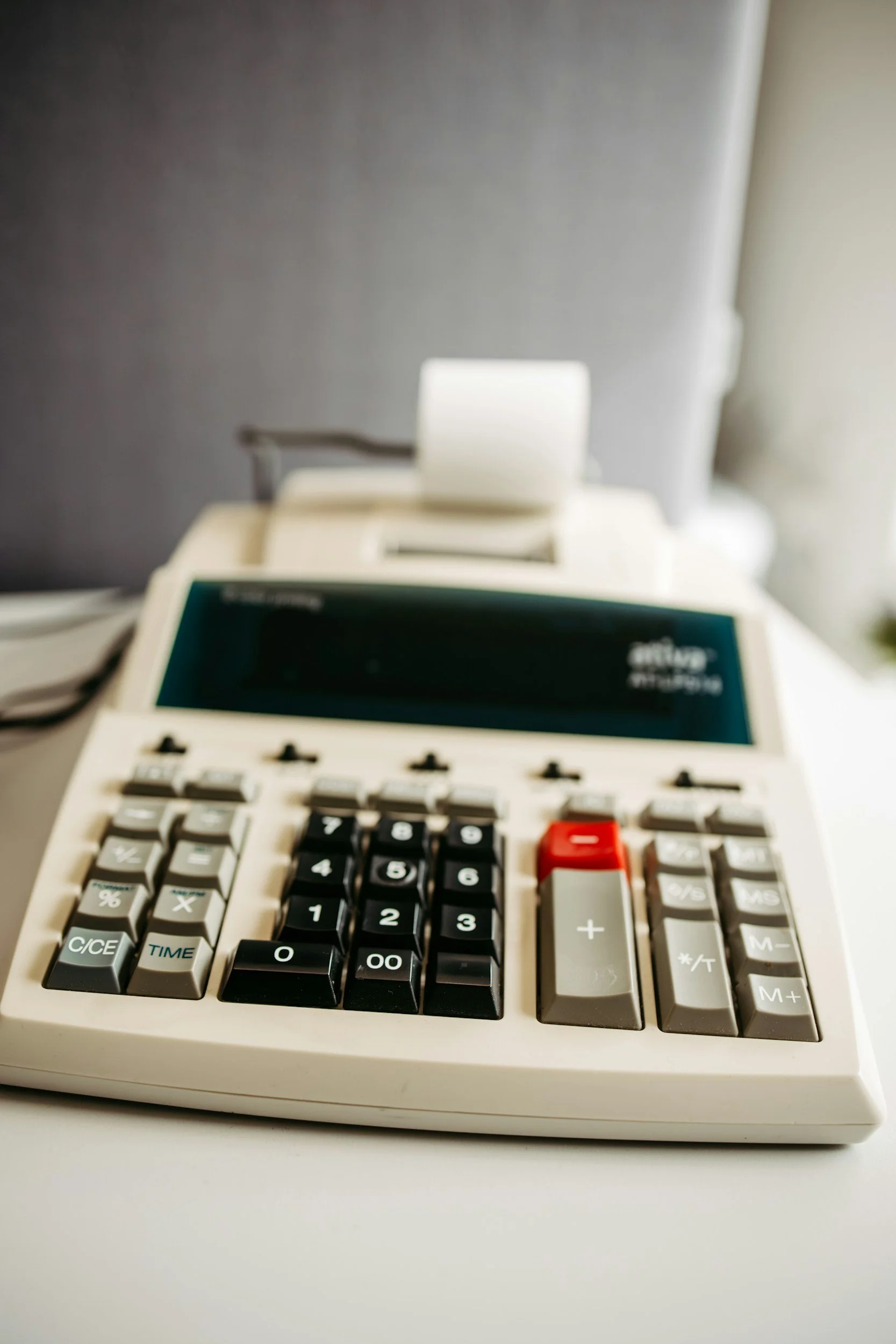 A close-up of a vintage calculator with black, gray, and red keys, placed on a white surface. The display is visible but turned off, and the background includes a gray wall and a blurred computer monitor.
