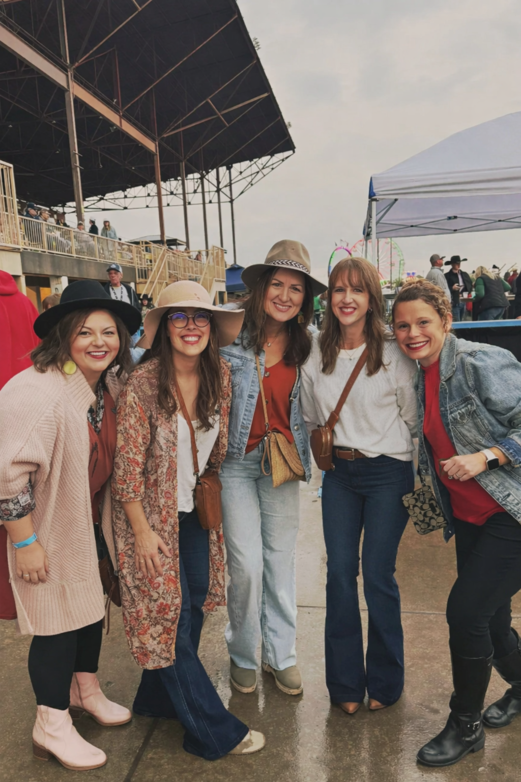 Five women smiling and posing together at an outdoor fair or festival, with a large covered stage and amusement rides in the background.