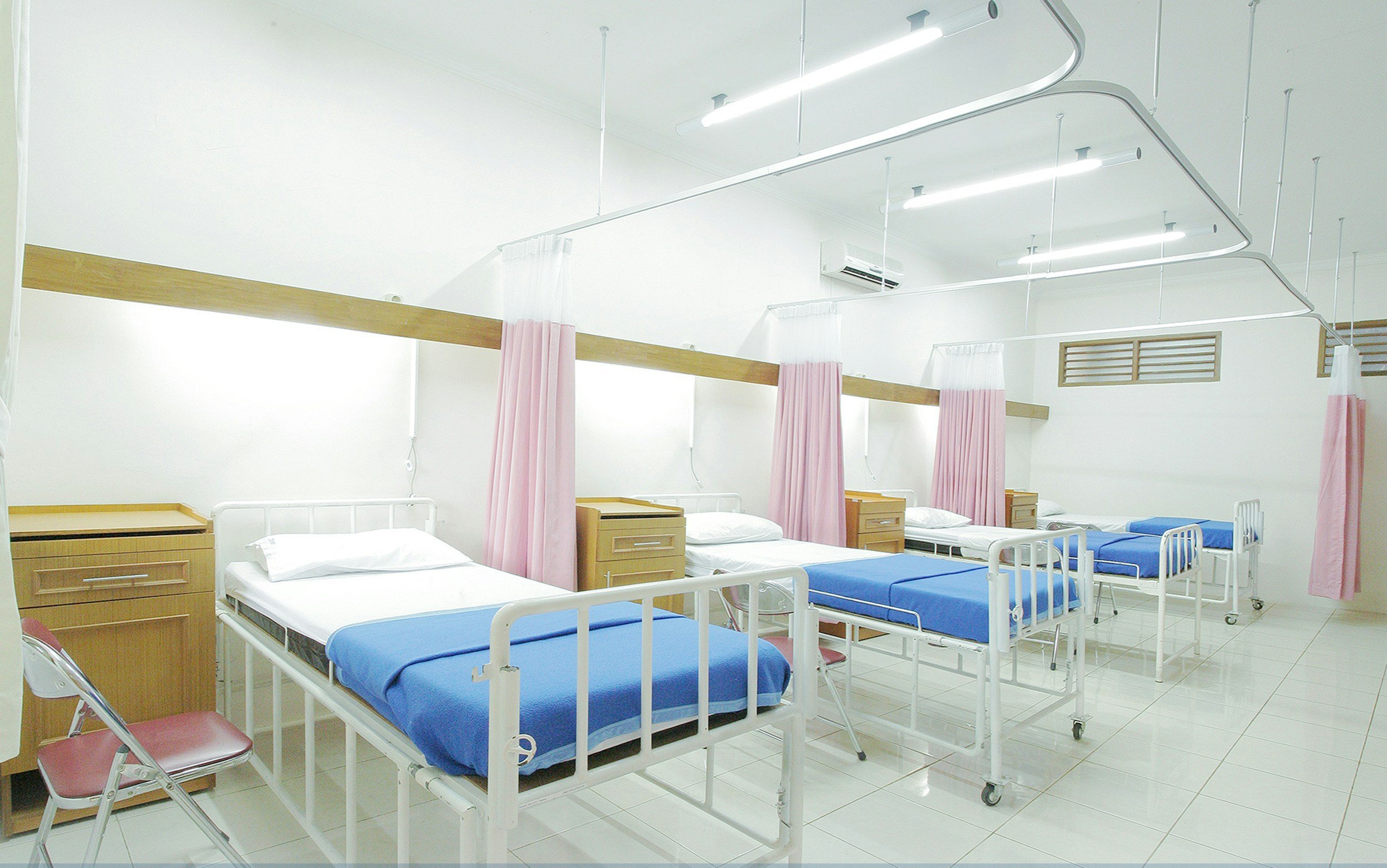 A hospital room with five beds, each separated by pink curtains, with white walls and a white tiled floor. Each bed has a headboard and blue or white bedding, with some beds on wheels. There are chairs beside some beds and overhead lights and a ventilation vent are visible on the ceiling.
