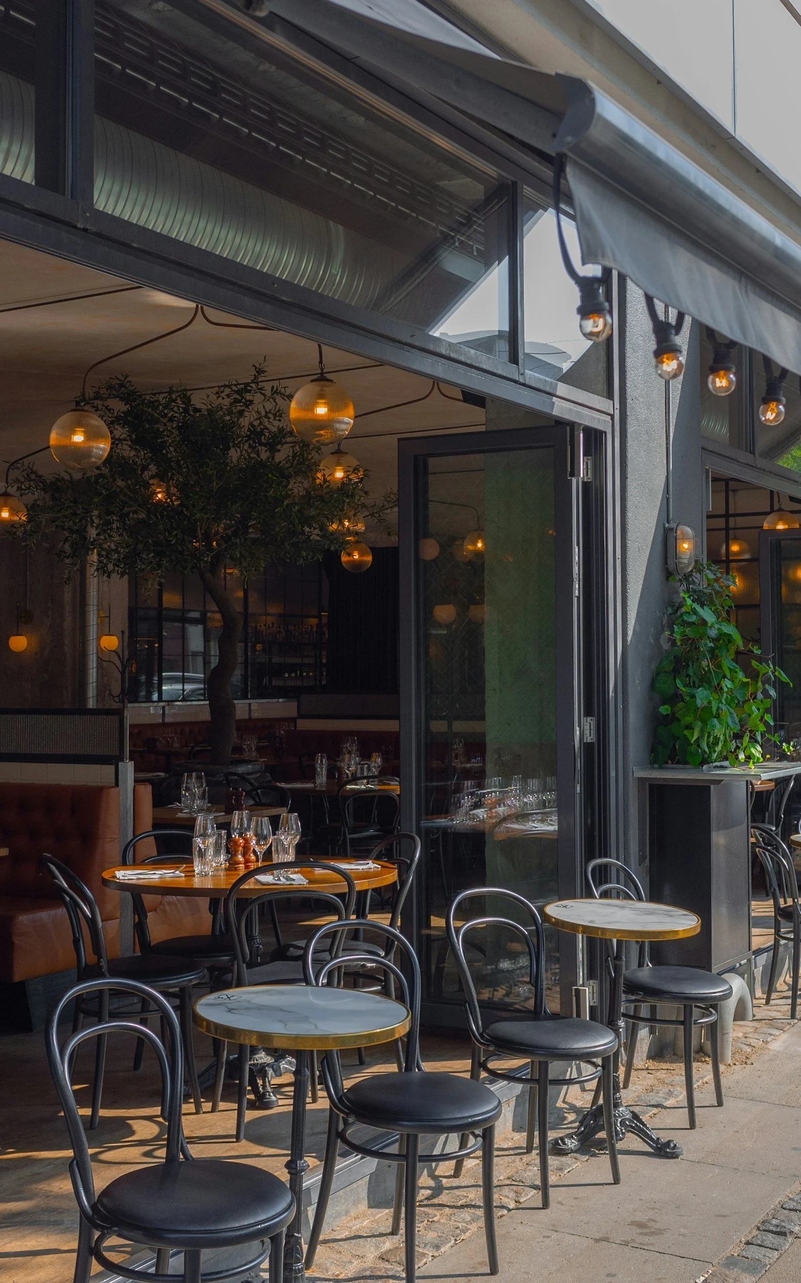 Outdoor view of a restaurant with tables and chairs, partially enclosed by glass walls, decorated with hanging spherical lights, plants, and outdoor string lights.