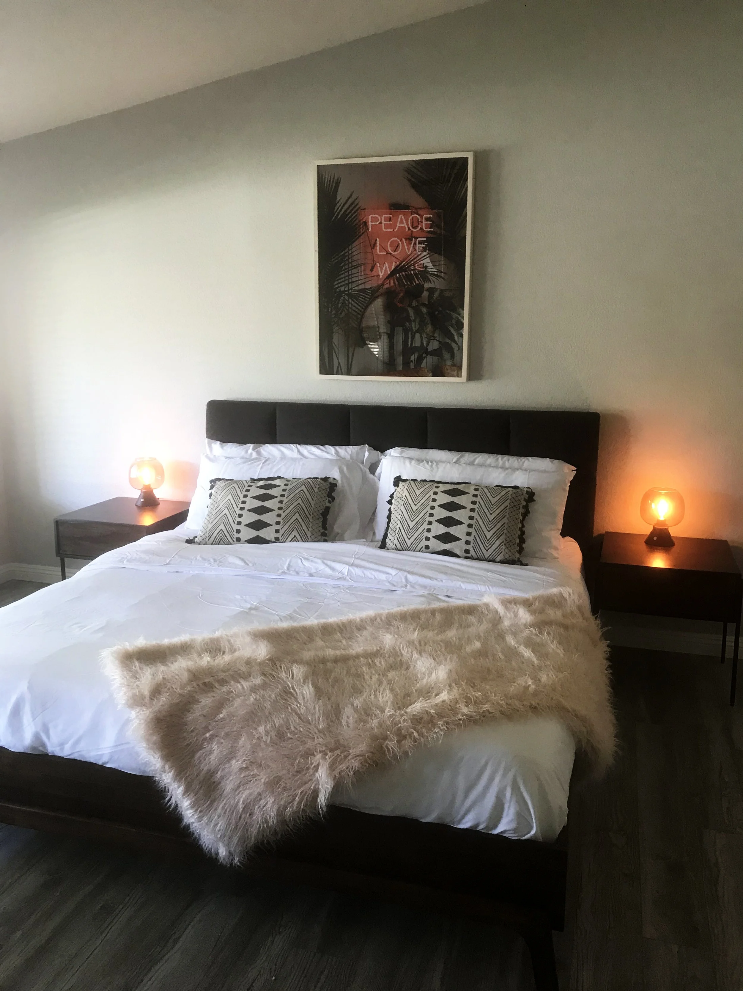 A bedroom with a bed, two nightstands with small lamps, a fur throw on the bed, two patterned pillows, and a framed artwork with palm leaves and the words 'Peace Love_War' on the wall behind the bed.