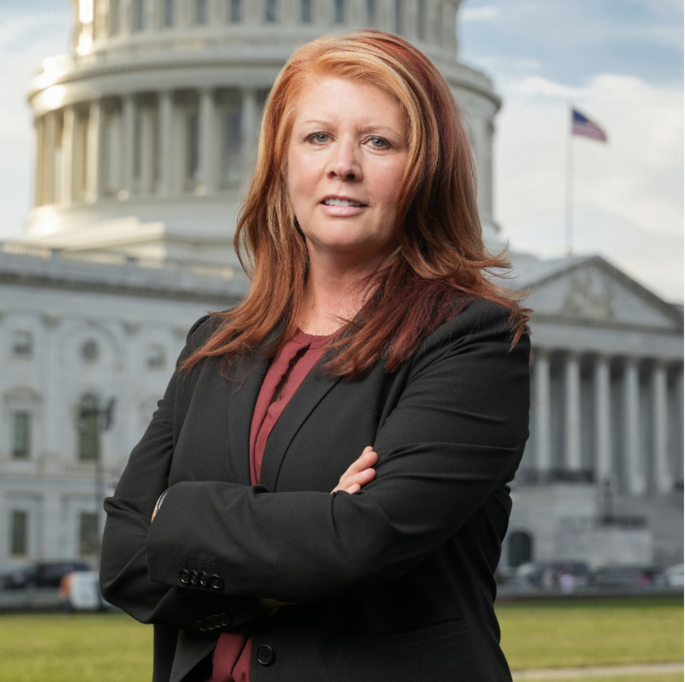 A woman with red hair wearing a black blazer and maroon shirt, standing in front of the US Capitol building with arms crossed.