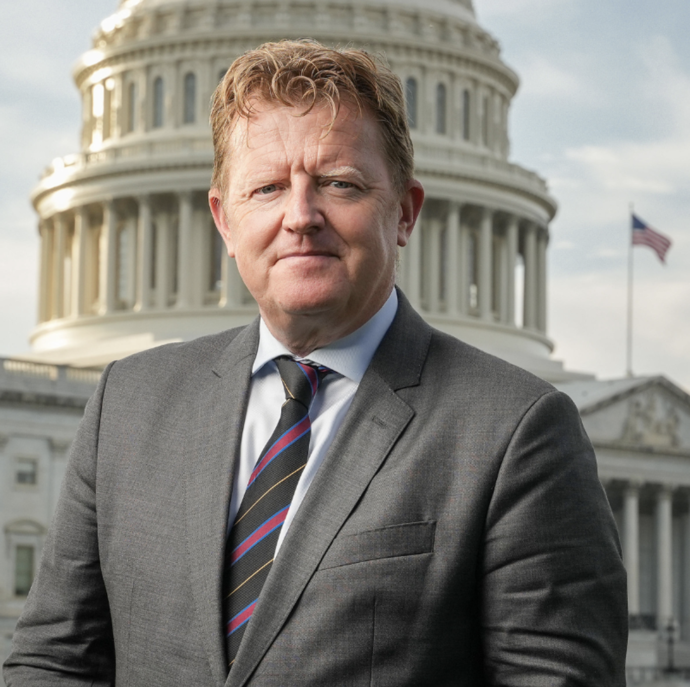 A man in a suit standing in front of the United States Capitol building with an American flag flying in the background.