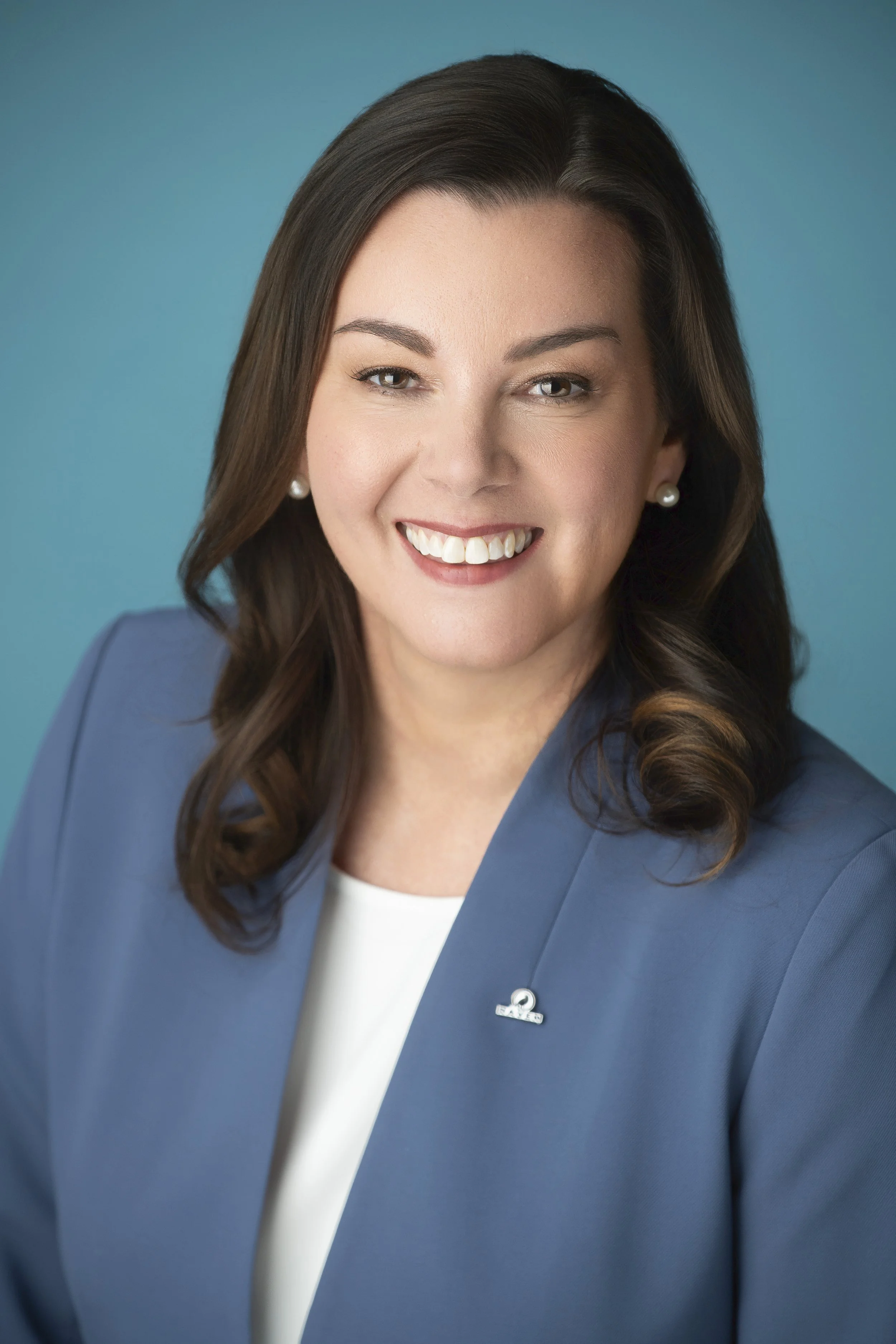A woman with brown hair smiling, wearing a navy blue blouse and silver earrings, posed against a gray background.