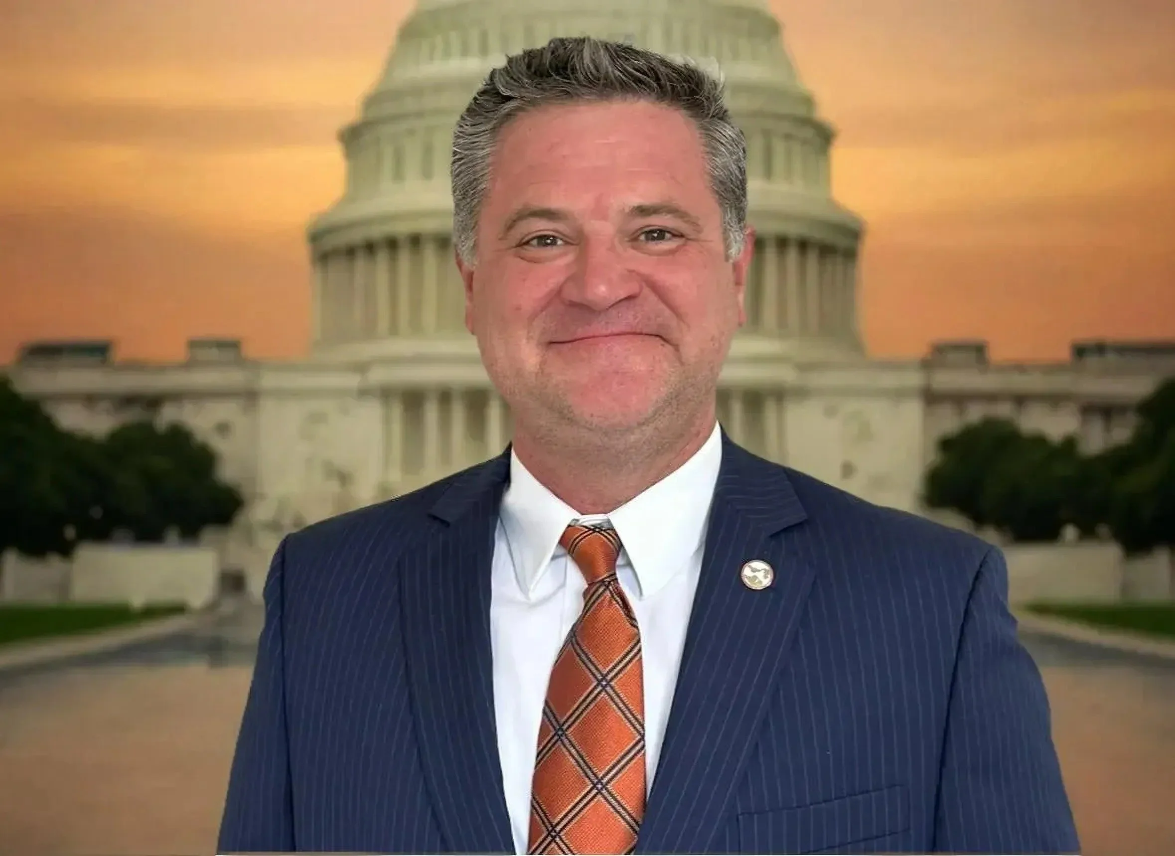 A man in a navy blue pinstripe suit with an orange and brown plaid tie smiling in front of a backdrop of the US Capitol building at sunset.