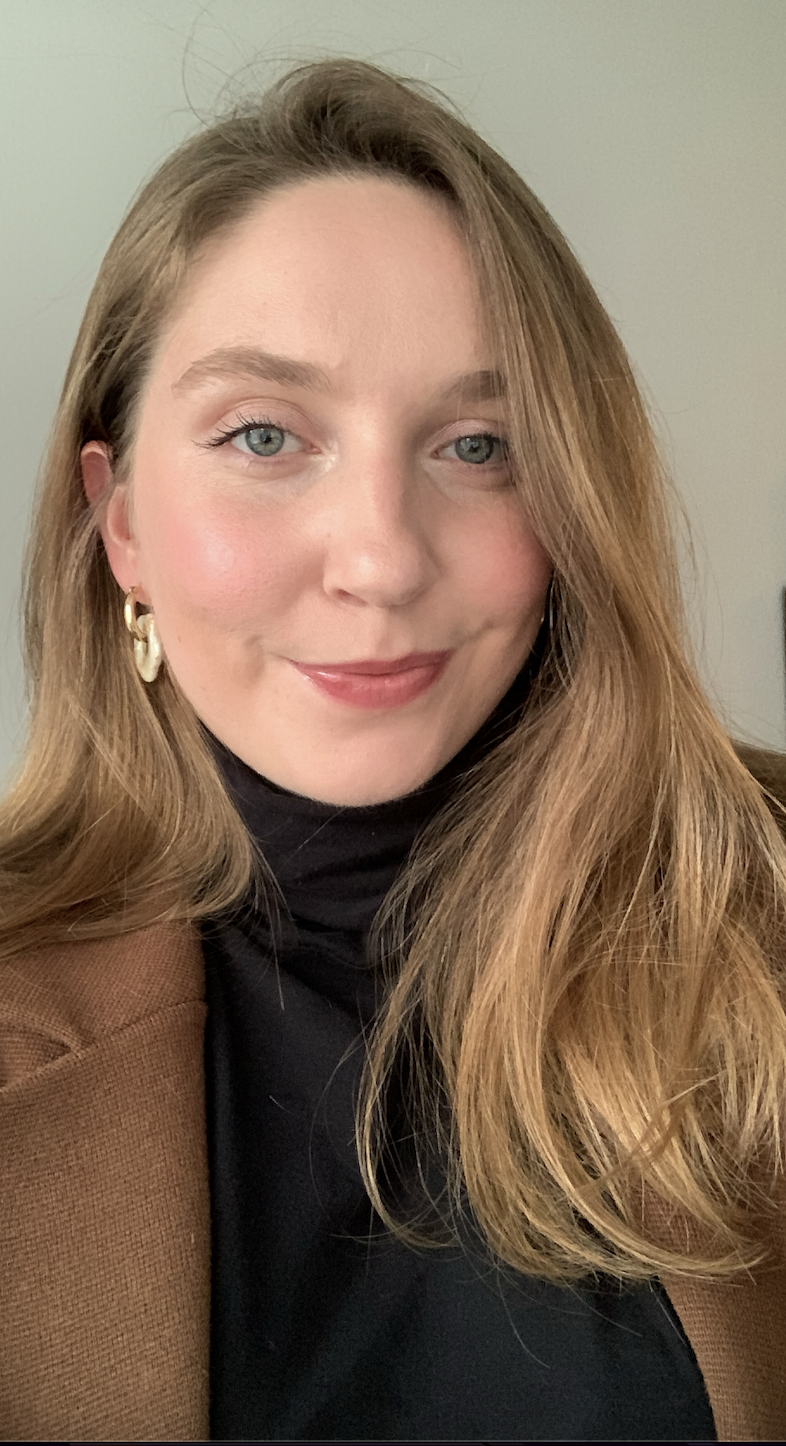 Close-up of a young woman with long, light brown hair and blue eyes, smiling gently, wearing a black turtleneck, brown blazer, and gold earrings.